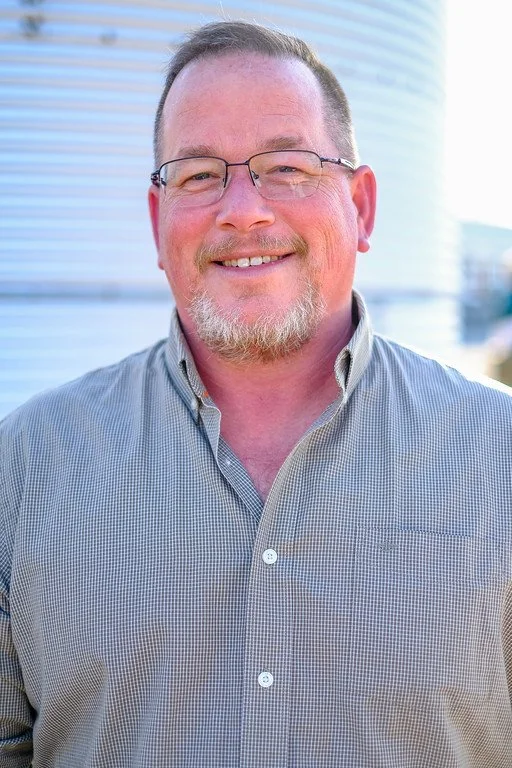 A middle-aged man with glasses, a beard, and short hair smiling outdoors, wearing a button-up shirt, with a modern building in the background.