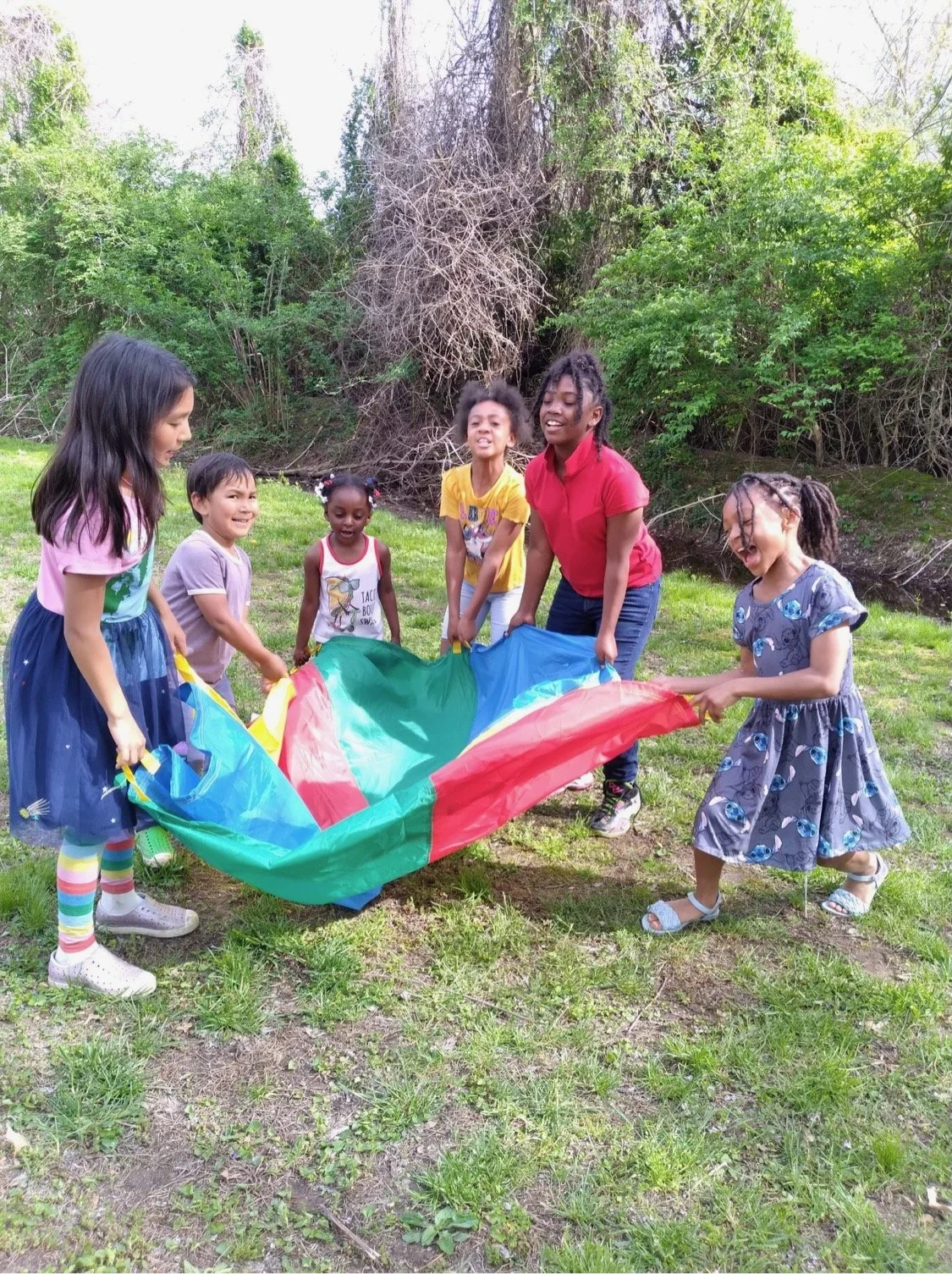 Children playing outdoors with a colorful parachute in a park surrounded by green trees.