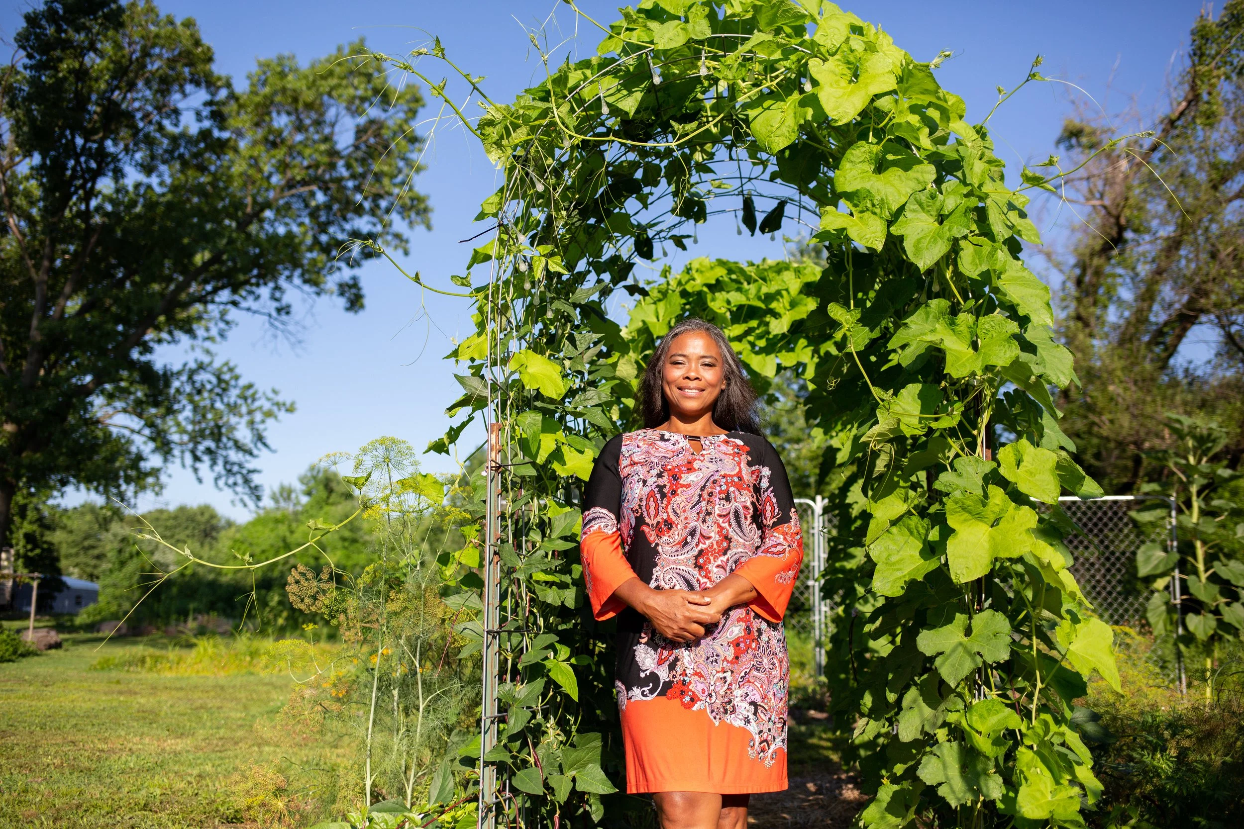 A woman standing under a large leafy arch made of green plants, smiling outdoors on a sunny day with trees and a fence in the background.