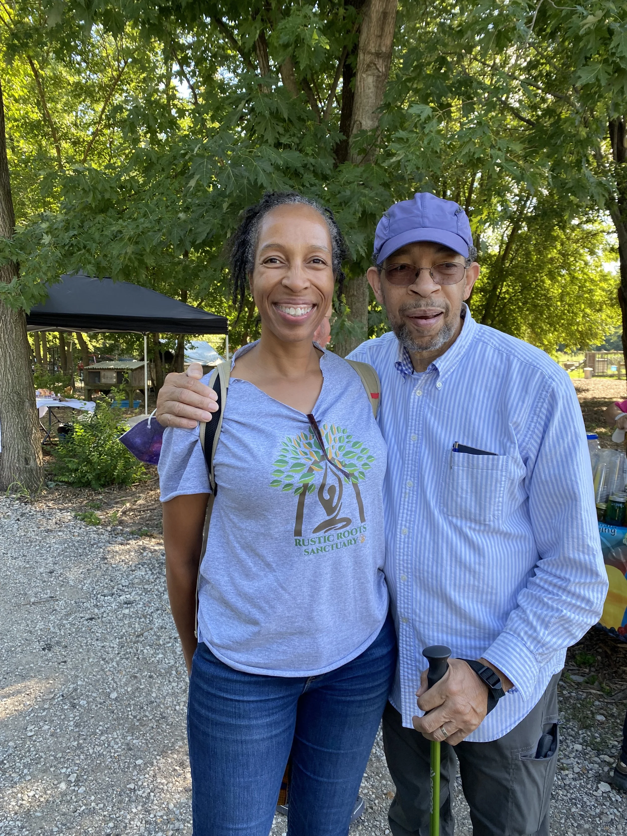 A smiling woman and a man with a walking cane outdoors under trees, with a canopy and other people in the background.