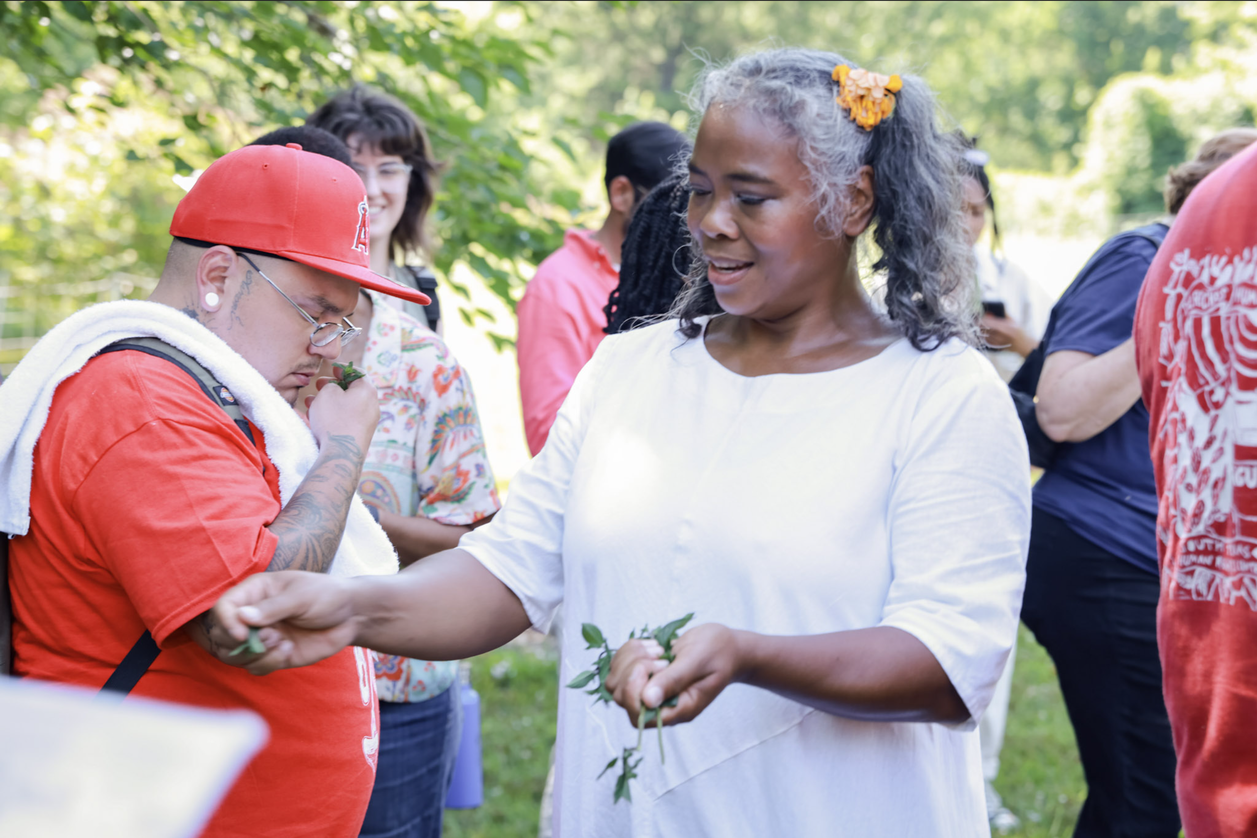 A group of diverse people outdoors, with one woman in a white shirt holding and displaying green leaves, others observing and participating in a nature activity.