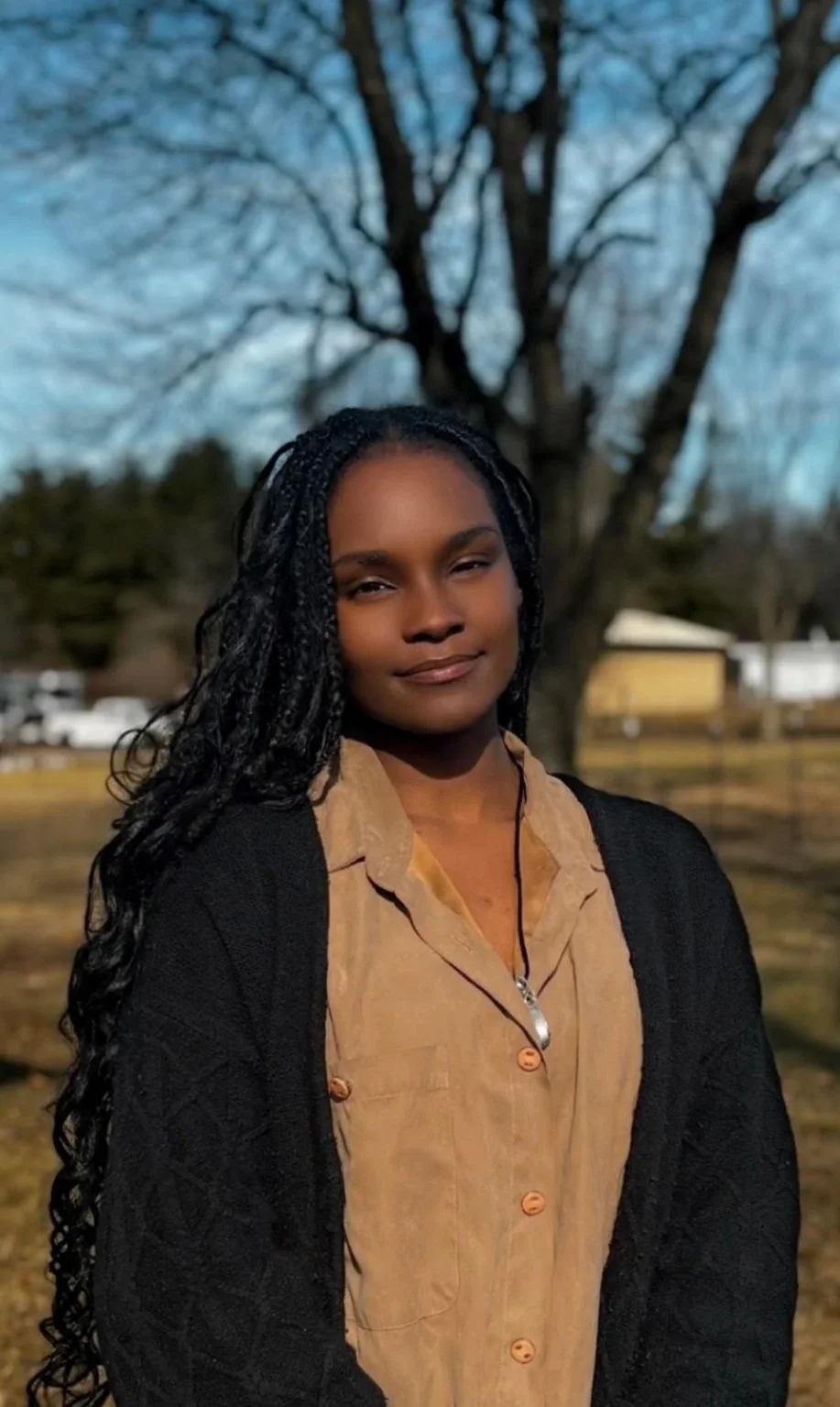 A woman with long curly black hair standing outdoors in front of a large tree with bare branches. She is wearing a tan button-up shirt, a black cardigan, and a necklace with a silver pendant. The background features a grassy area, some buildings, and a clear blue sky.