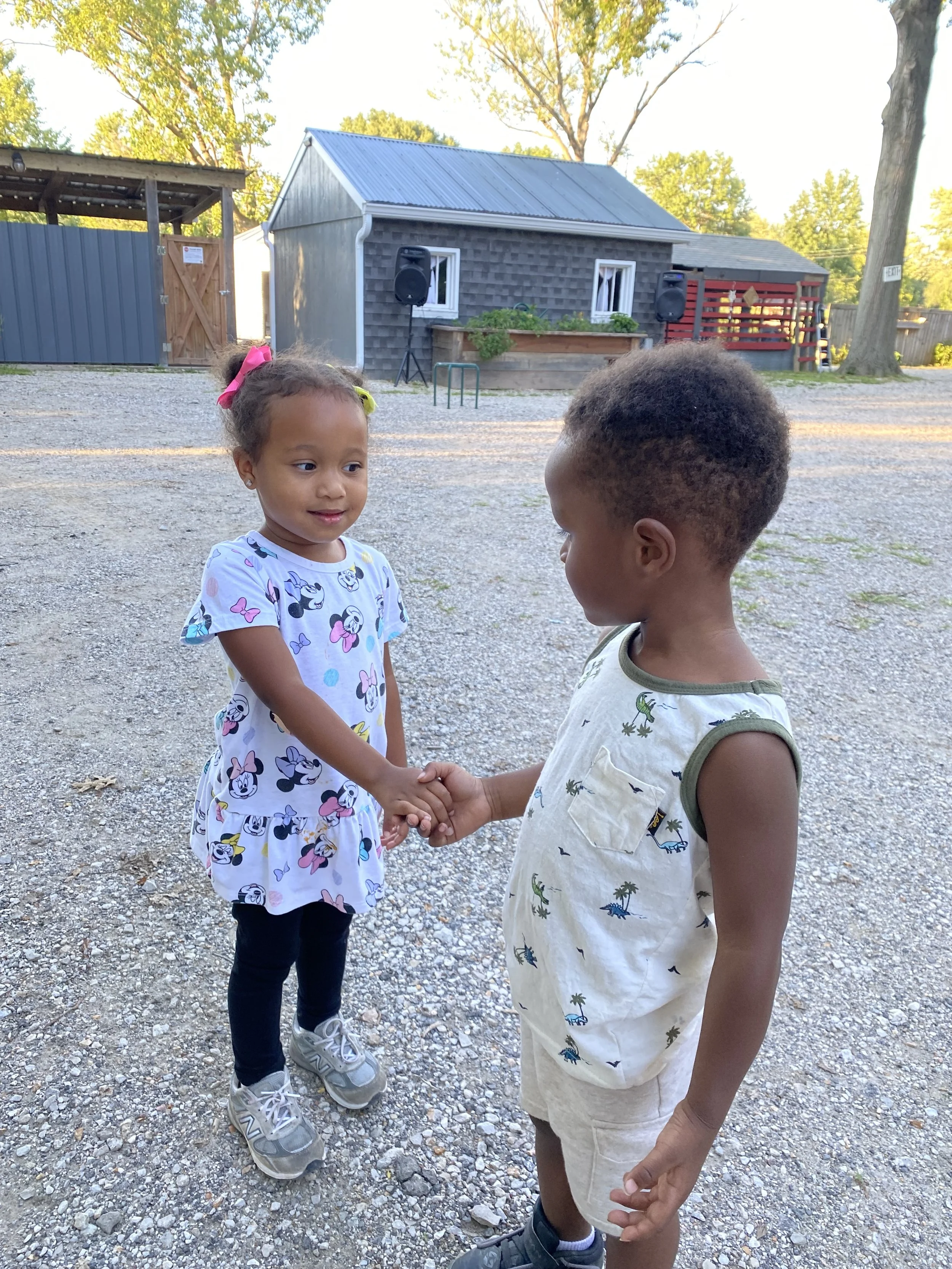 A young girl and boy holding hands outside in a gravel area, with small buildings, trees, and a speaker system in the background.