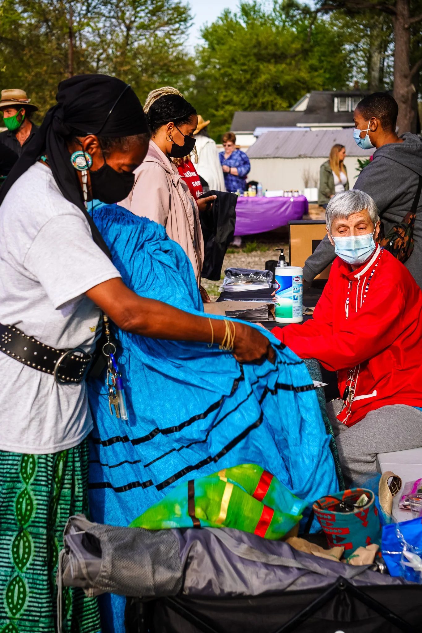 People wearing masks at an outdoor event, with some standing in line and others seated at tables, surrounded by trees and tents in the background.