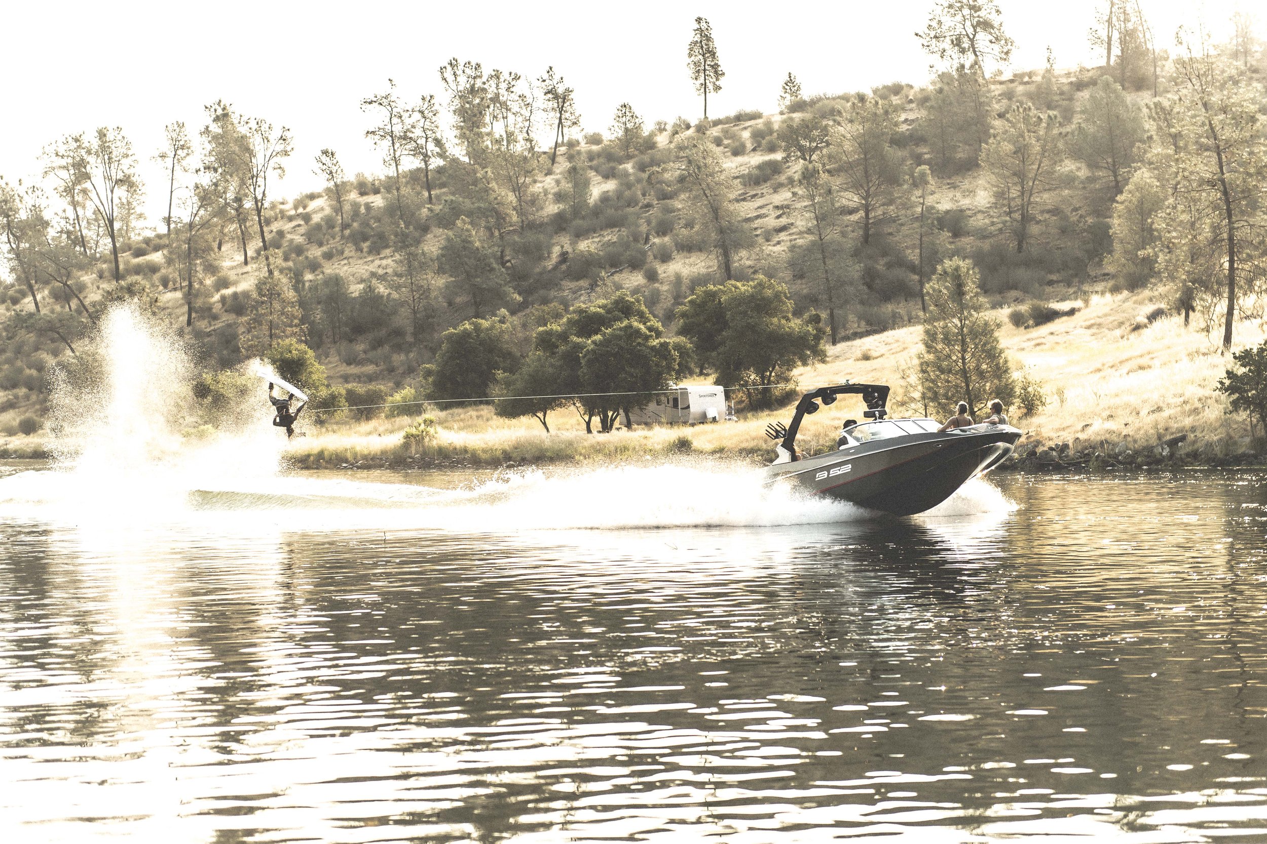 A person wakeboarding behind a motorboat on a lake with a hillside and trees in the background during sunset.