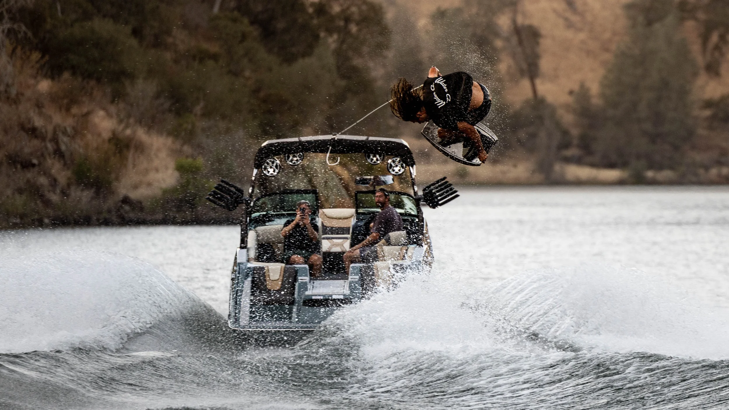 A person wakesurfing in the water, mid-air after launching off the boat, with two people on the boat watching and taking photos, on a serene lake with a forested shoreline in the background.