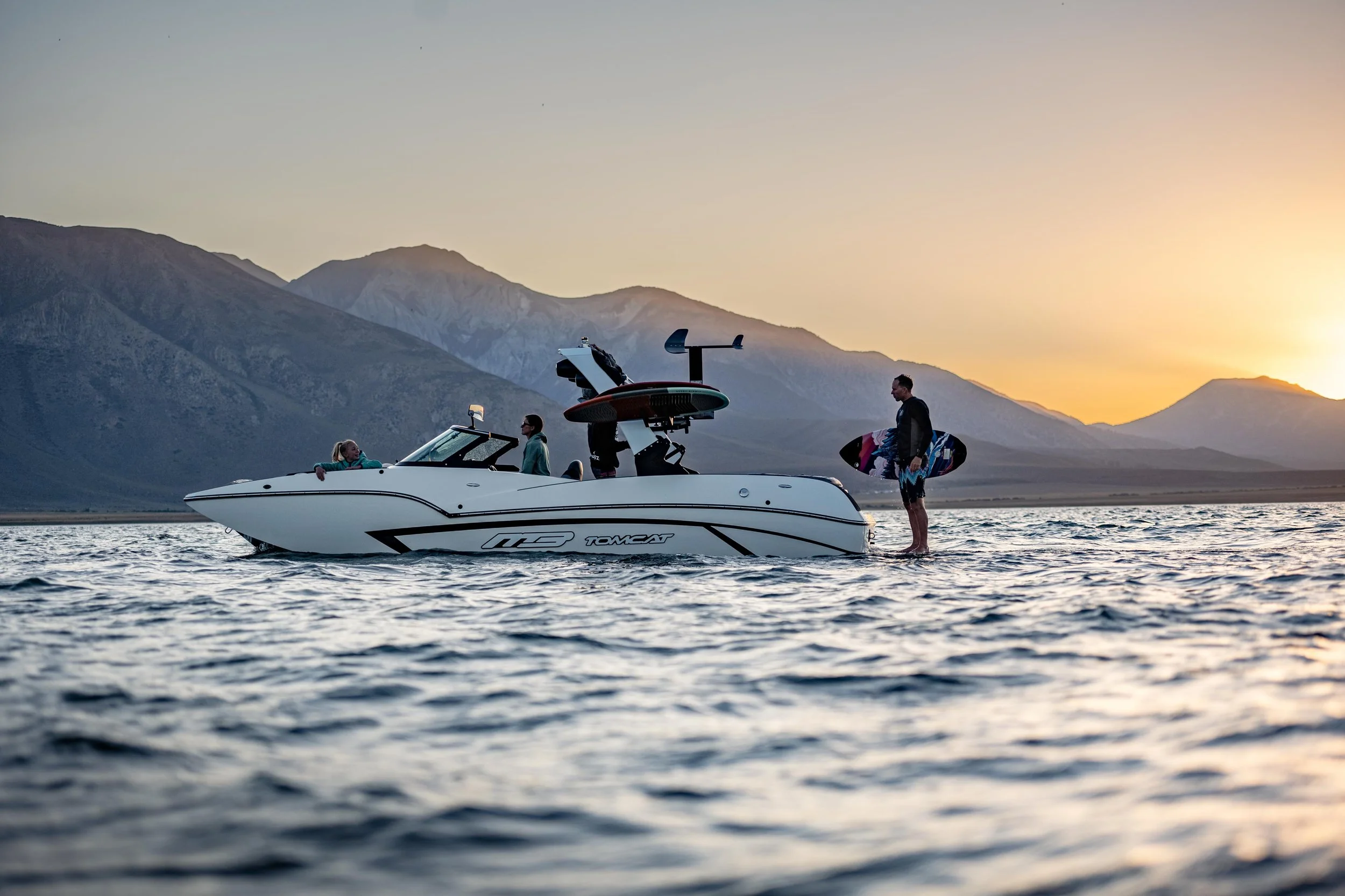A boat on water with three people on board and one person standing in water holding a surfboard, mountains and sunset in the background.
