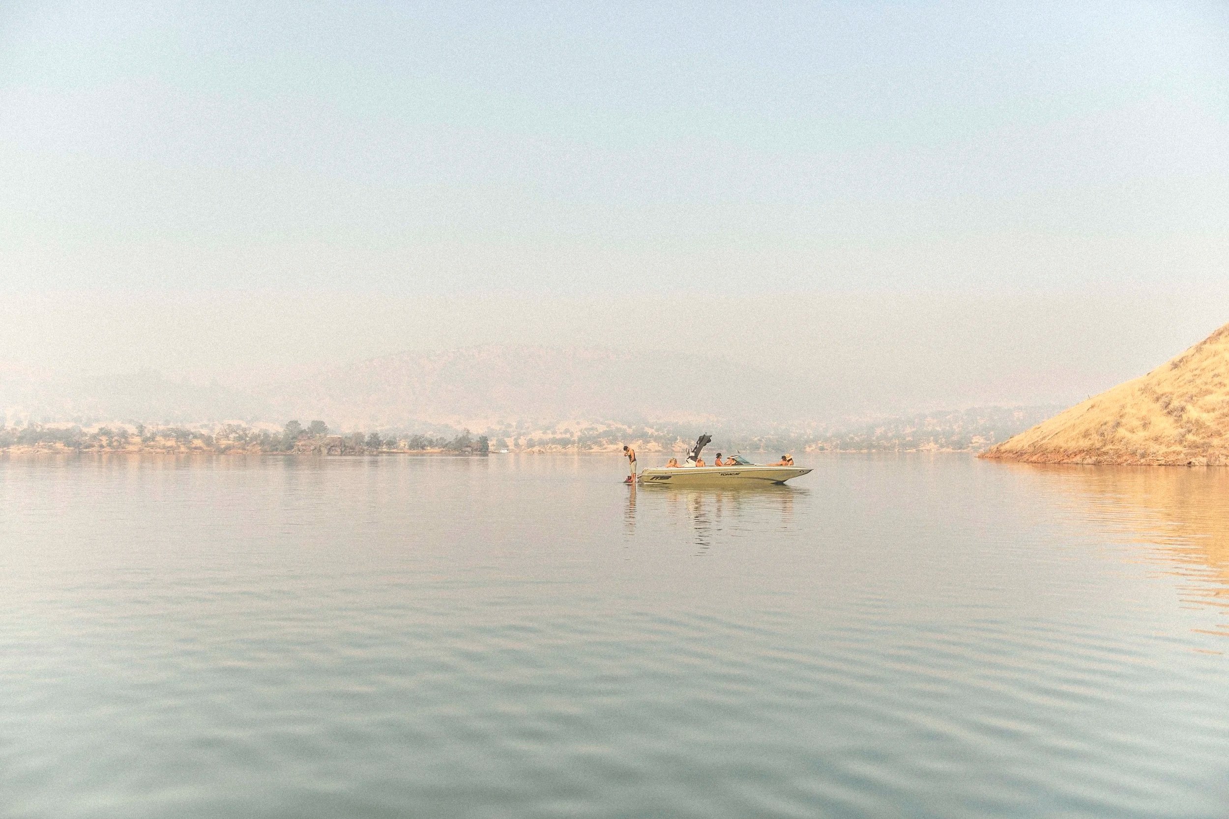People boating and standing on a paddleboard on a calm lake with a hilly shoreline in the background.