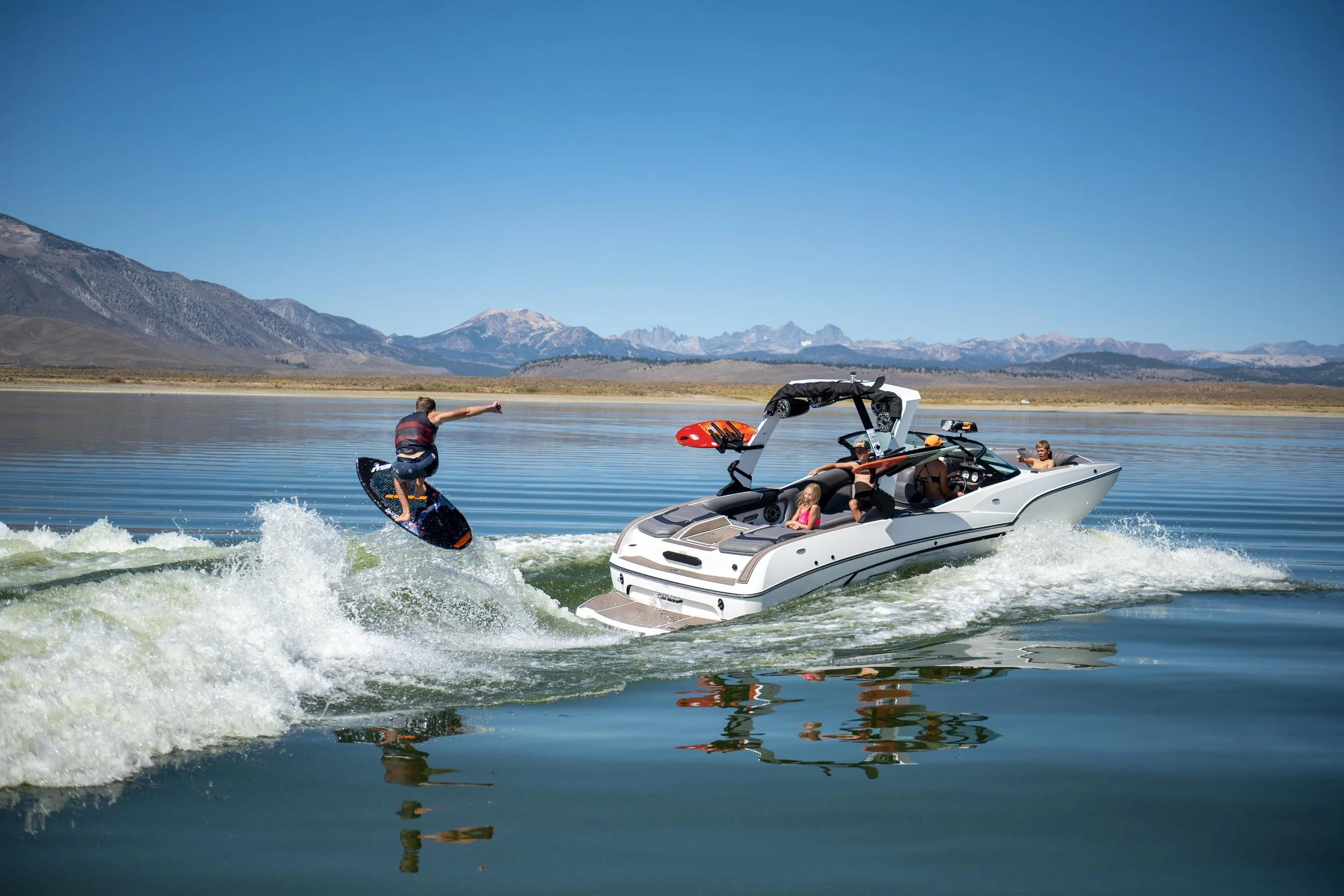 A person wakeboarding on a lake with a boat in the background, mountains and blue sky in the distance.