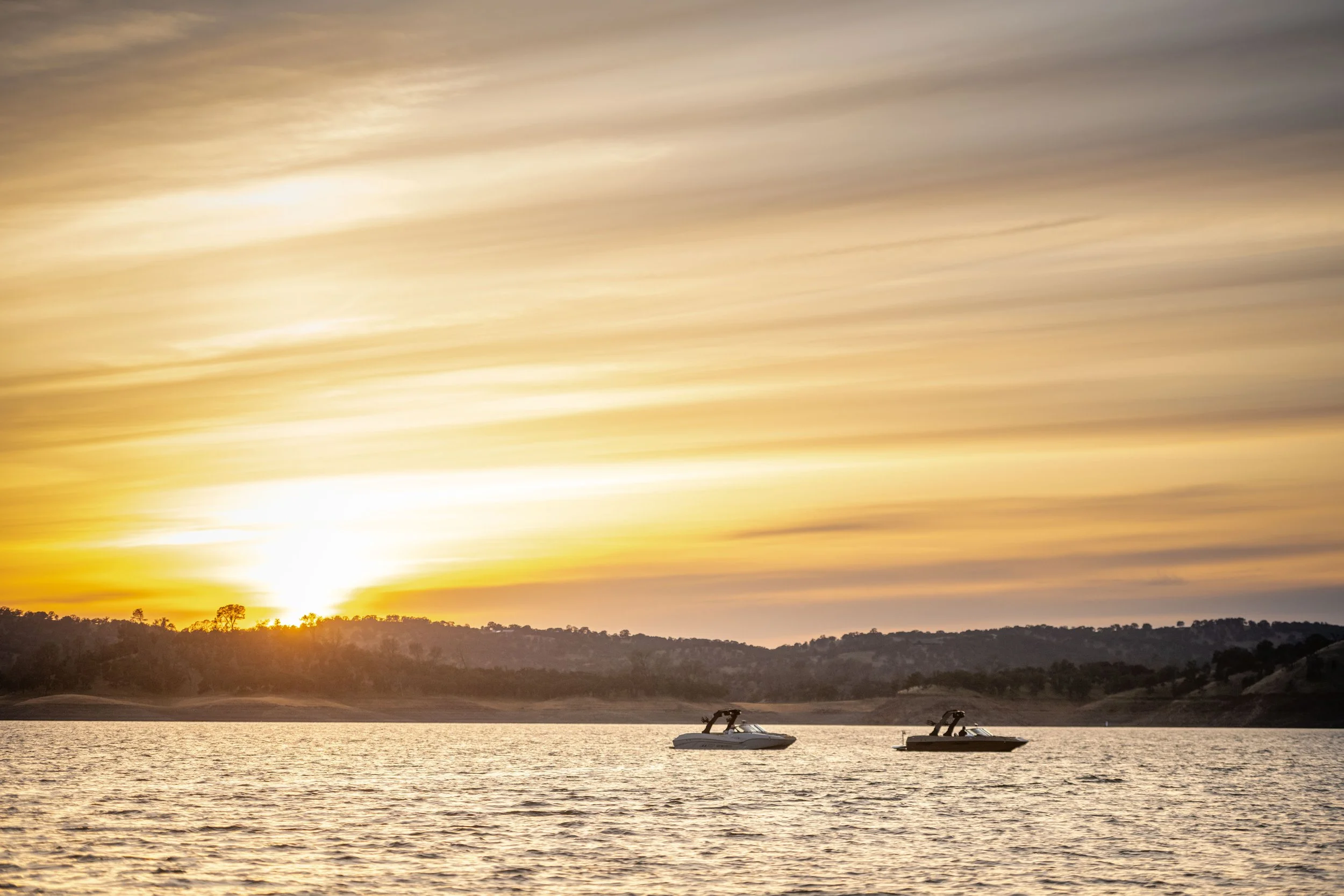Two boats floating on a lake at sunset with hills and trees in the background and colorful sky.