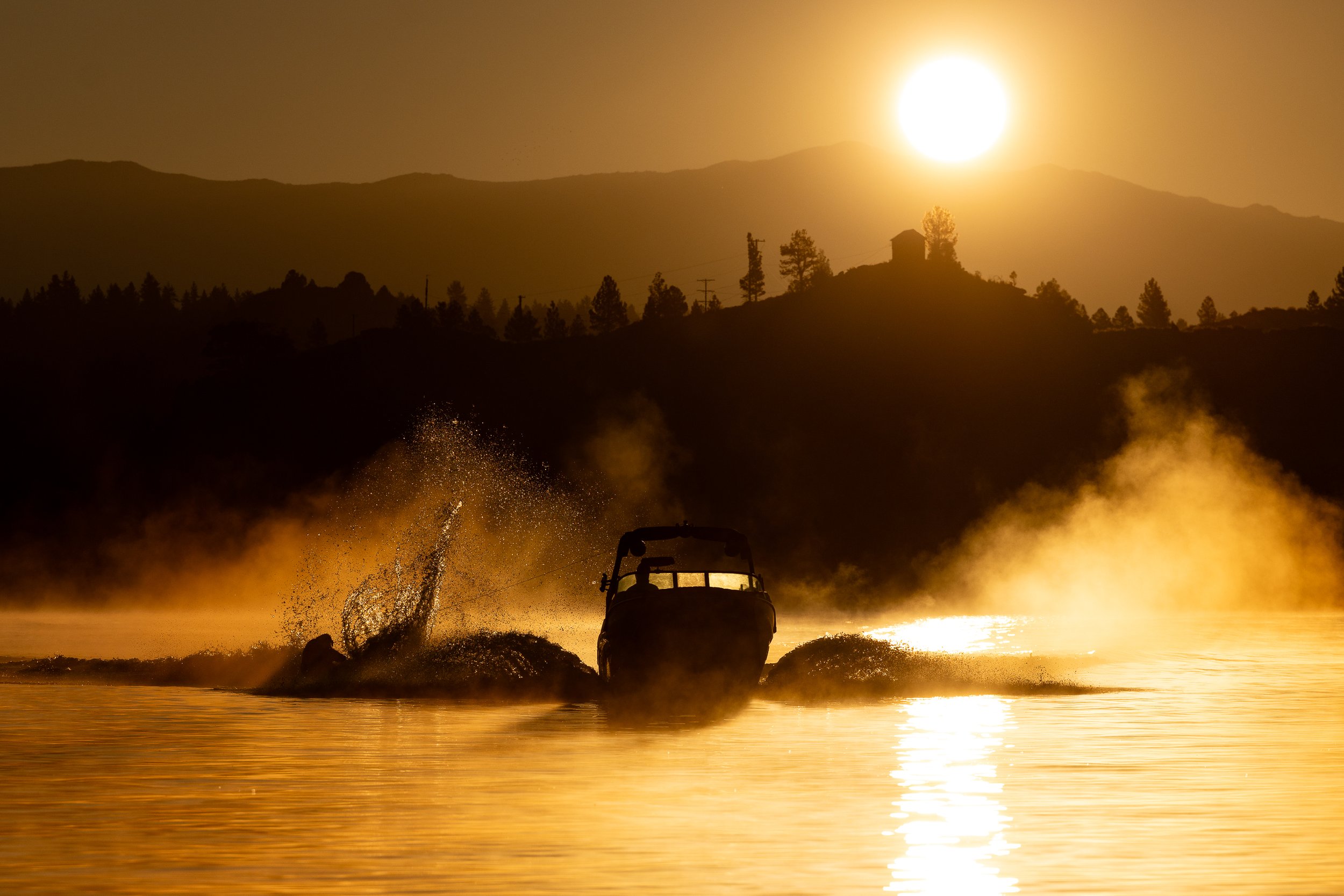 A boat speeding across a lake at sunset, creating a splash with the sun setting behind distant mountains and silhouetted trees.