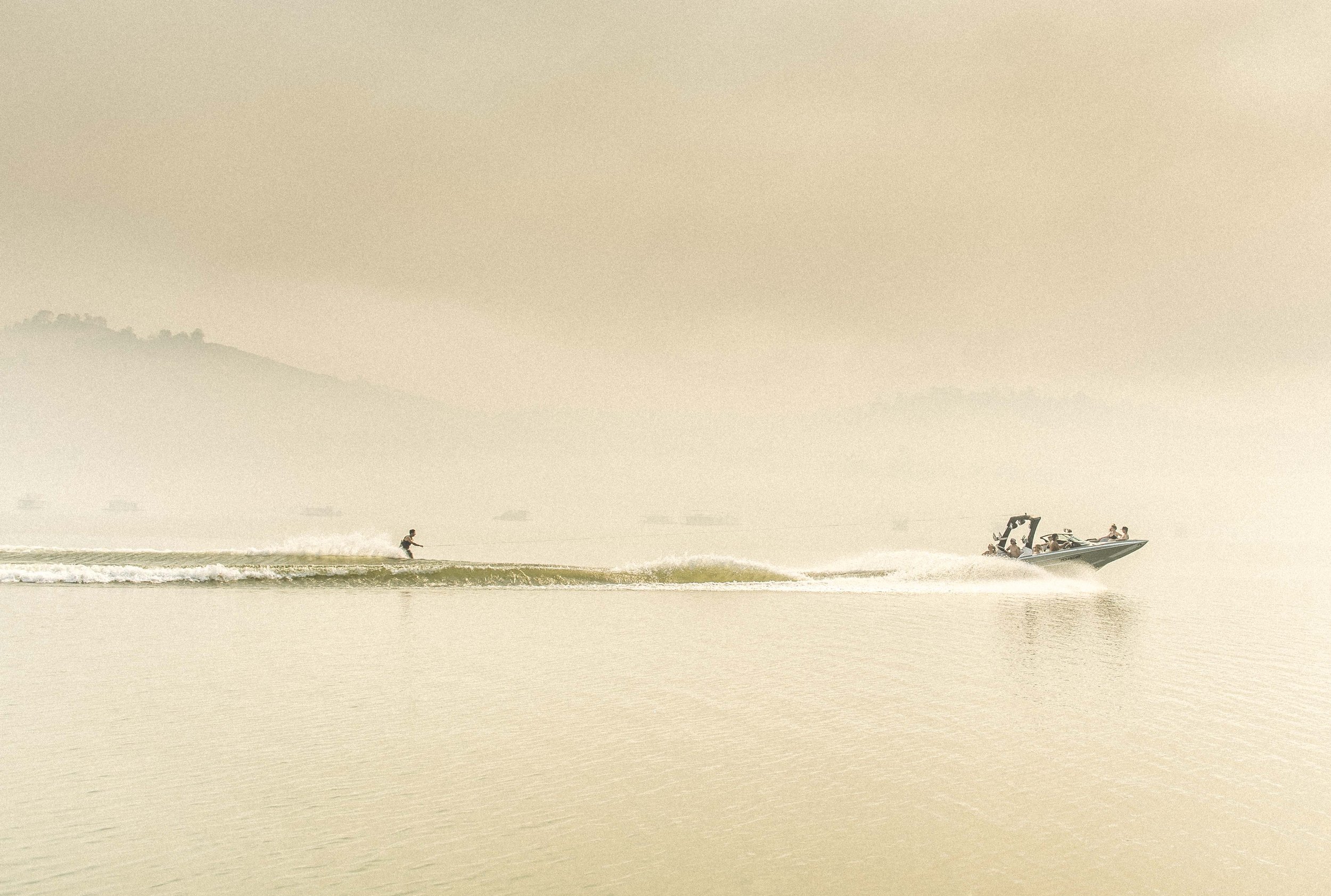 A person wakeboarding on a calm body of water, being pulled by a speedboat with a curved windshield and a canopy, with a hazy mountainous background.