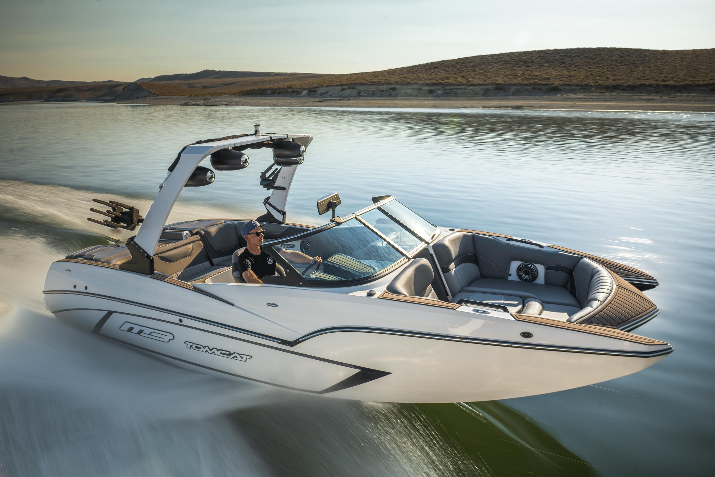 A white and gray motorboat cruising on a calm lake with a man driving, surrounded by a landscape of rolling hills and water.