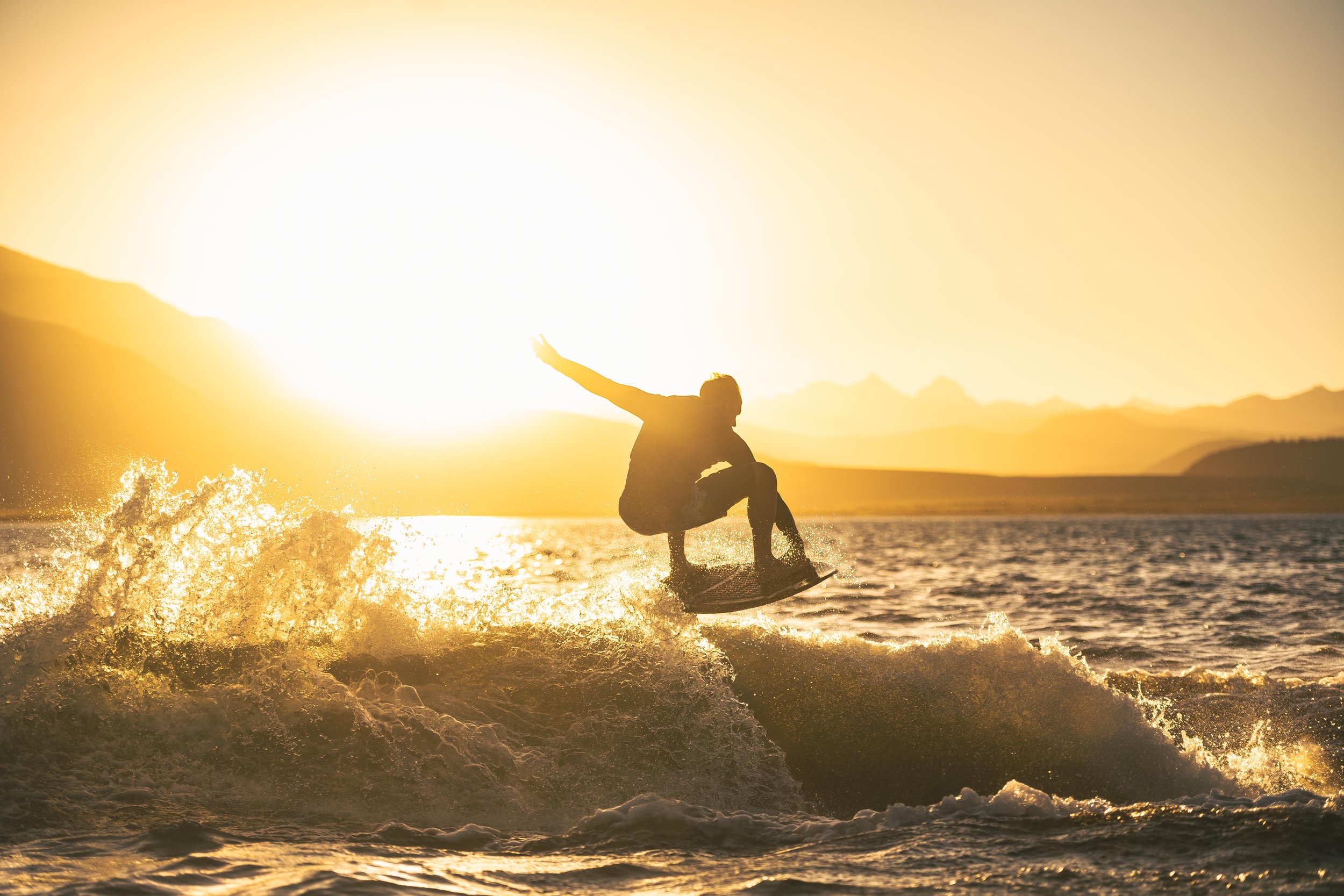 Silhouette of a person surfing on a wave during sunset with mountains in the background.