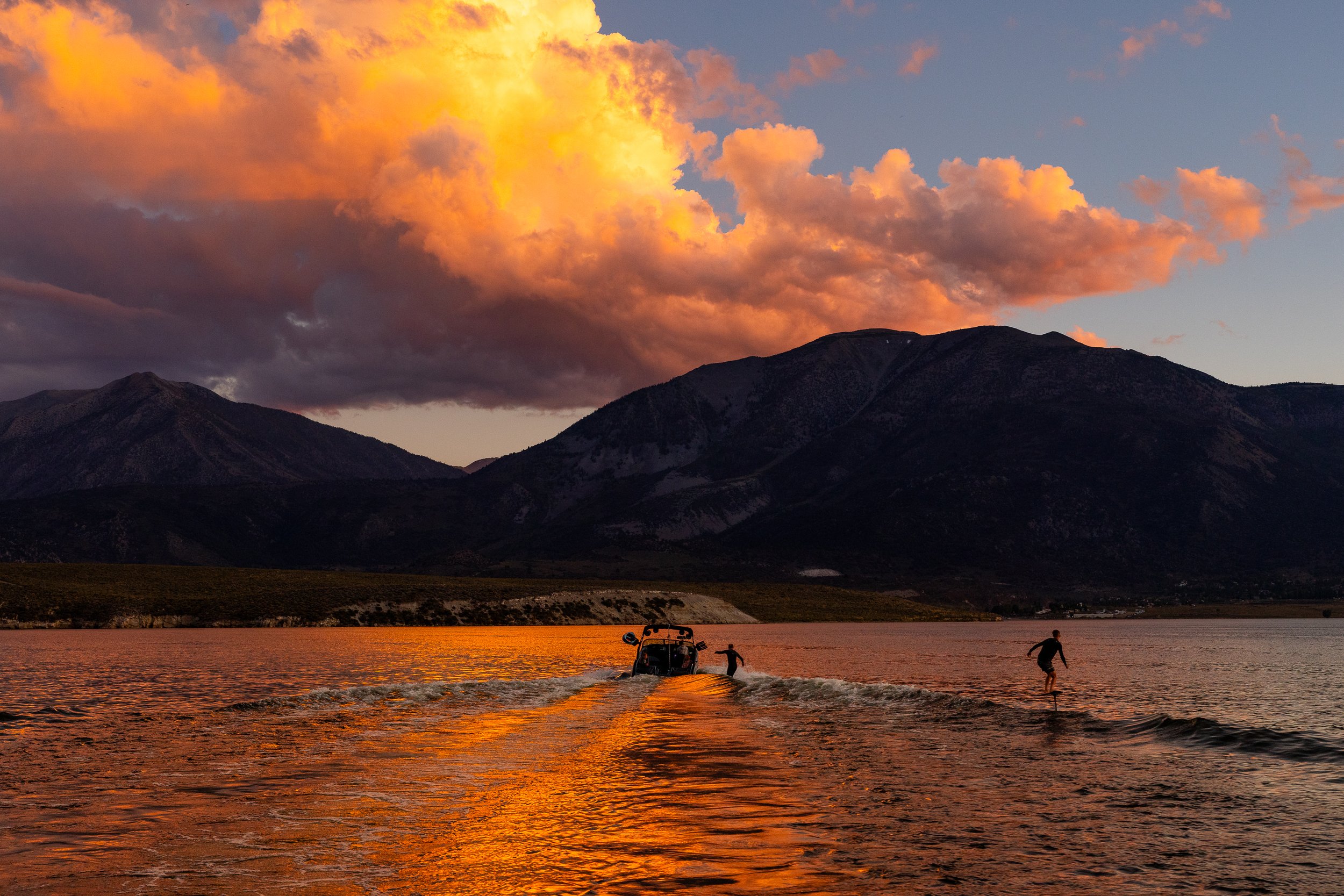People wakeboarding on a lake at sunset with mountains in the background and a colorful cloud-filled sky.