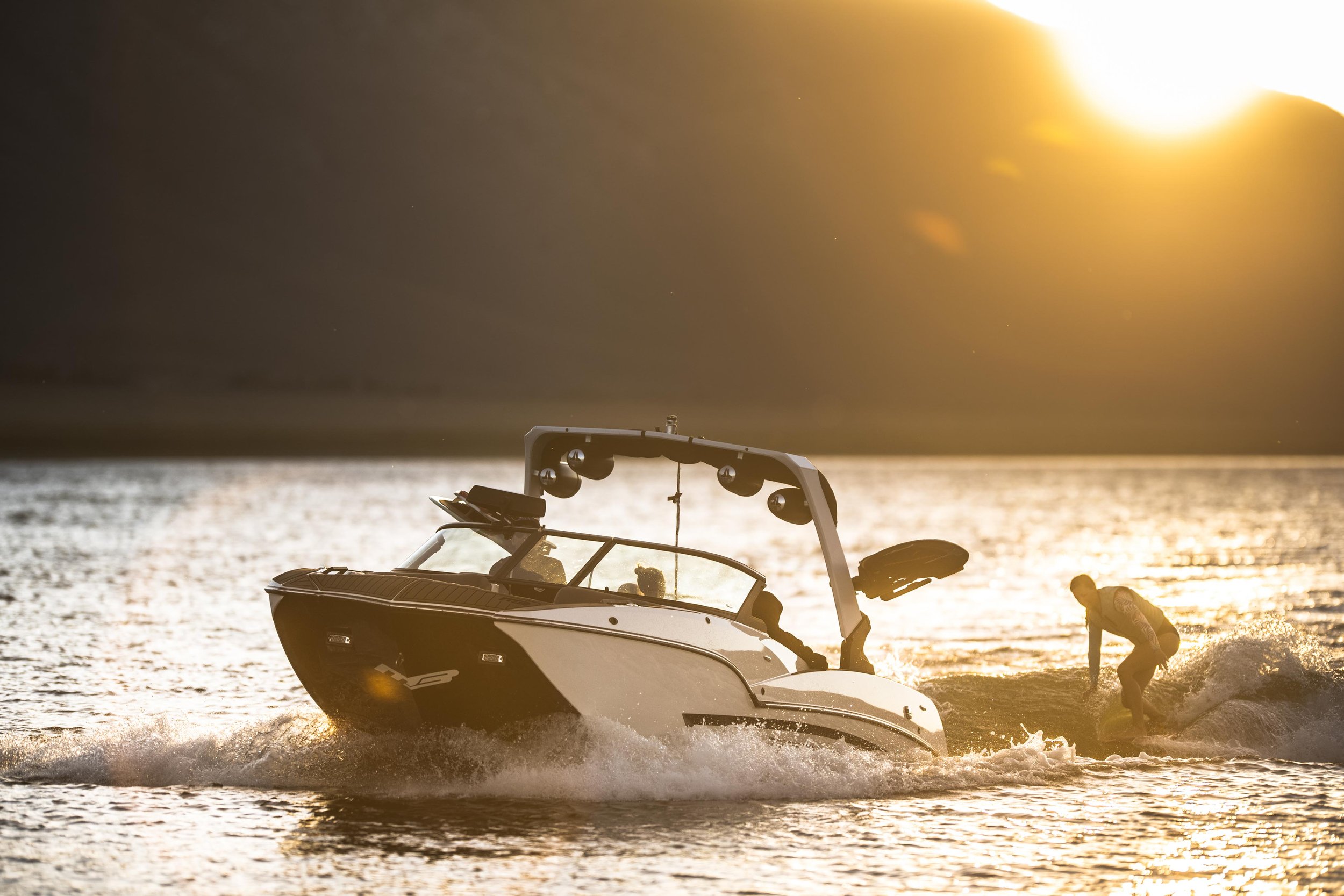 A person wakes on a surfing board on a boat in the water during sunset.