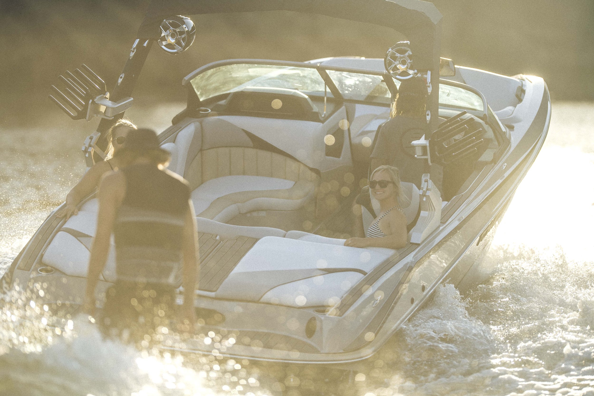 People enjoying a boat ride on a lake during sunset, with one woman wearing sunglasses and a striped top sitting comfortably at the front, and another person standing near the rear of the boat.