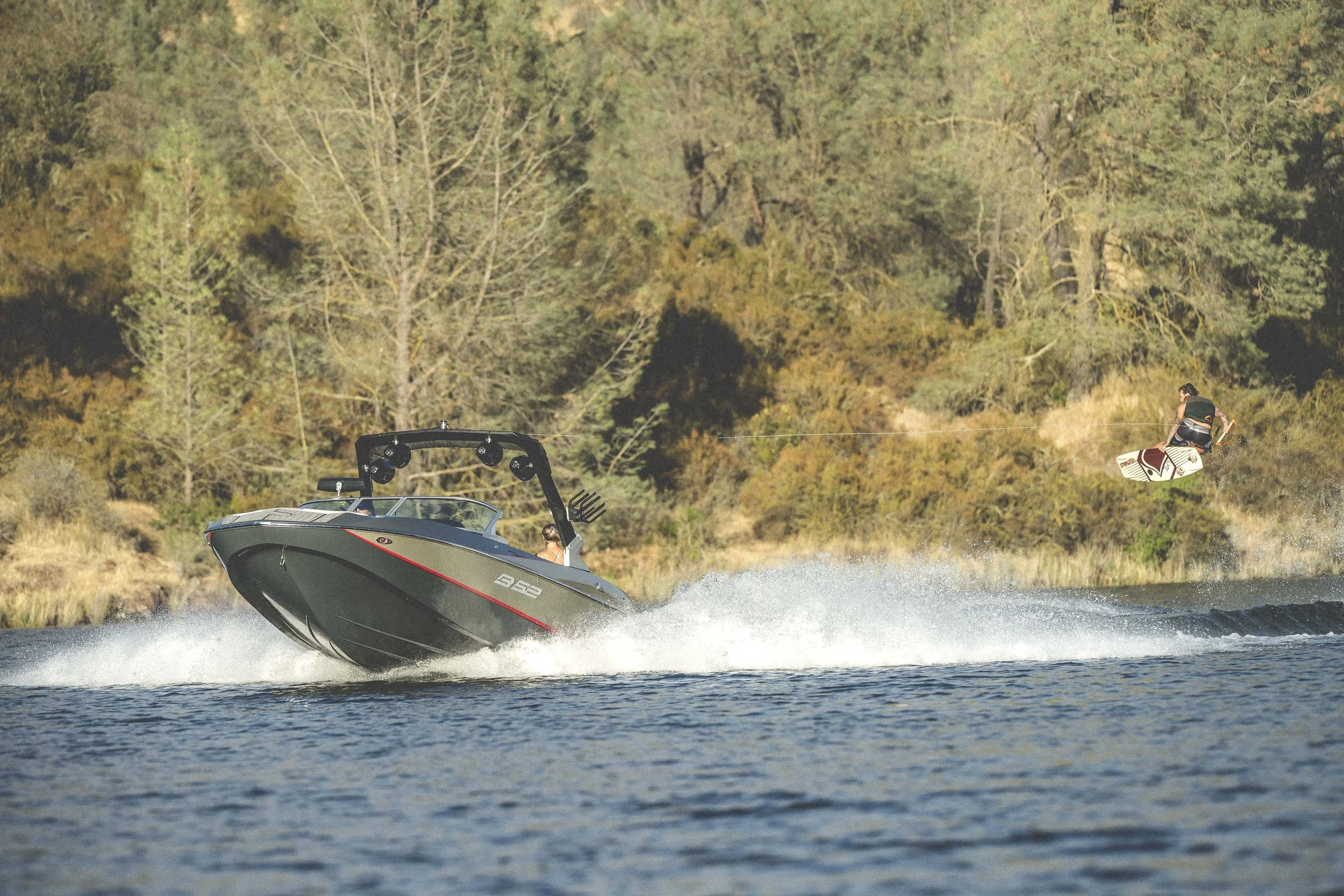 A speedboat on a lake with a wakeboarder being pulled behind, performing an aerial trick, with trees and shrubs along the shoreline in the background.