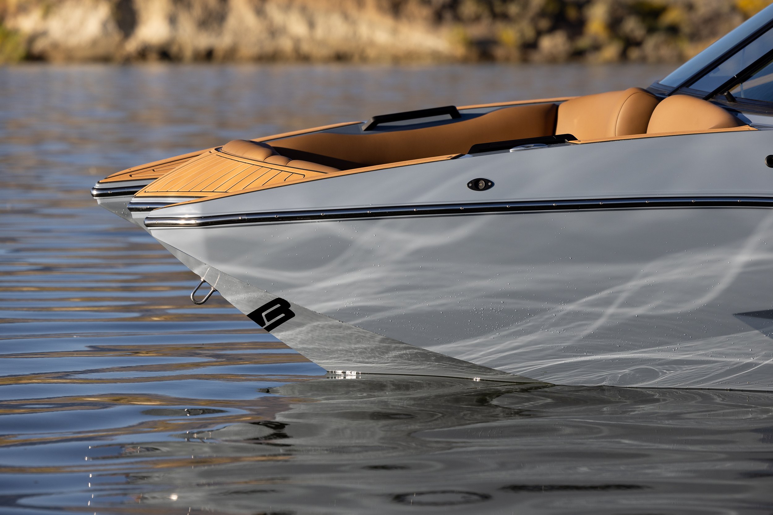 Close-up of a sleek, modern motorboat with a gray hull and tan interior, floating on calm water with a rocky shoreline in the background.