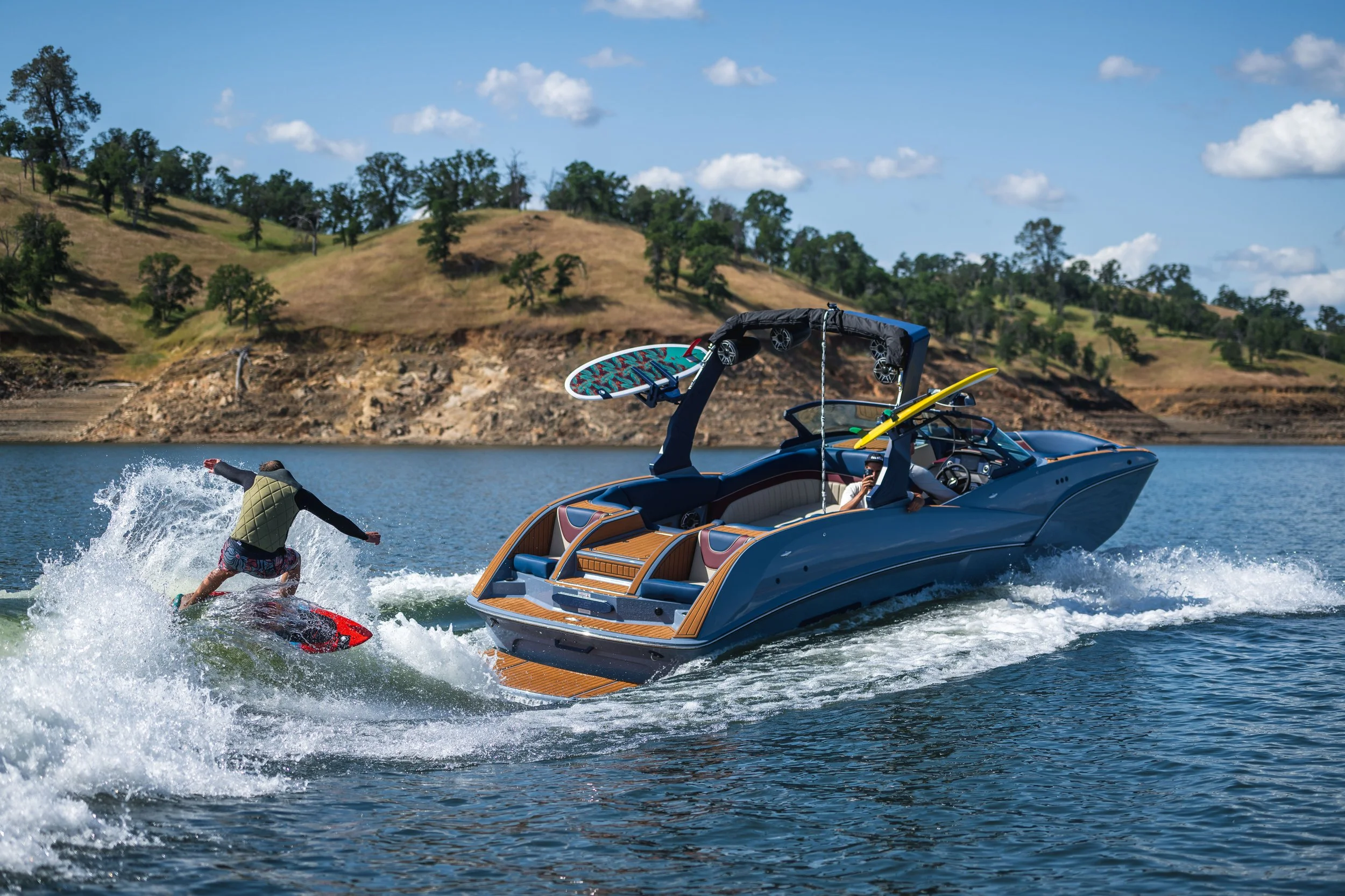 A person wakesurfing behind a boat on a lake with hills and trees in the background.