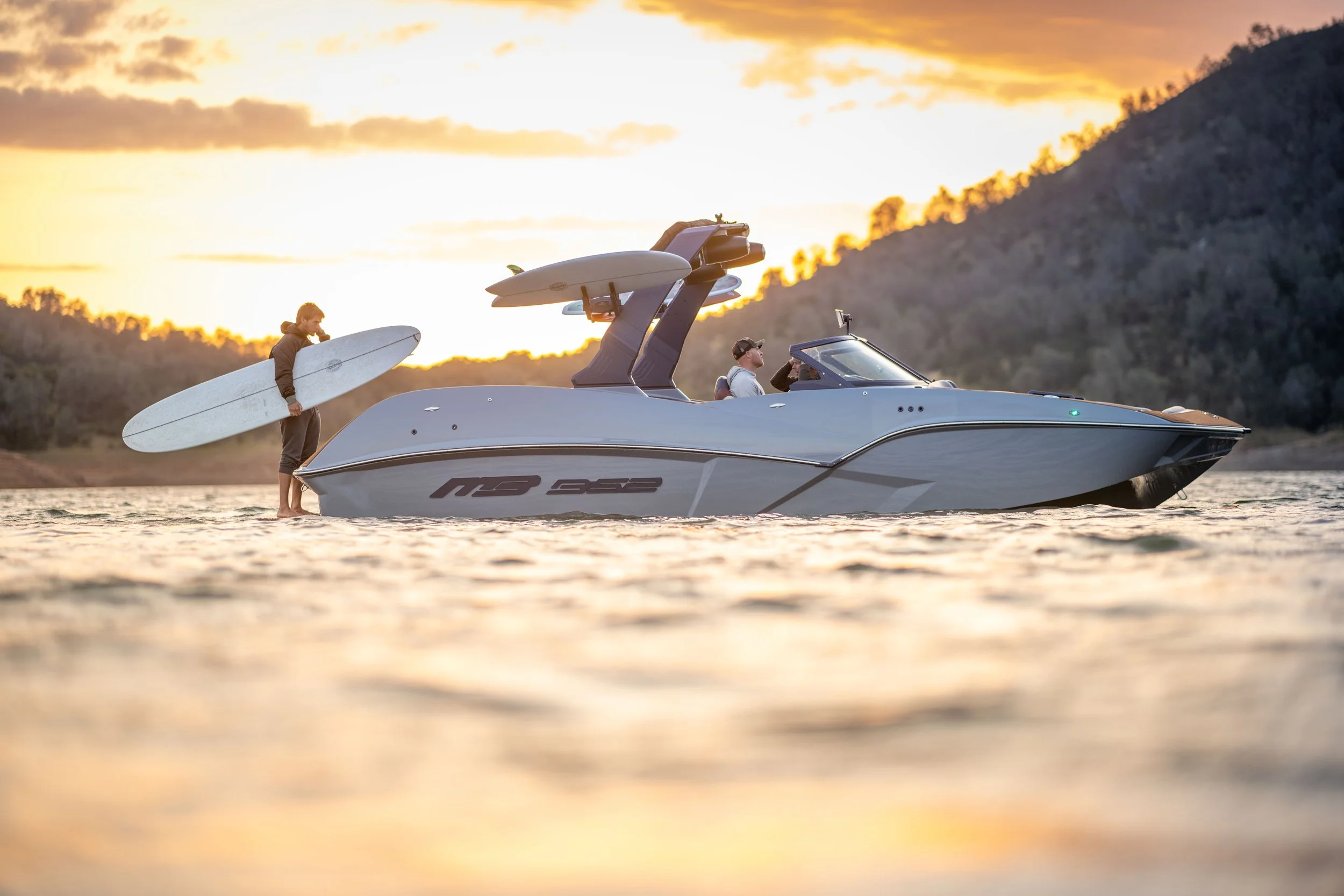 A boat with two people on a body of water at sunset, one person is in the boat steering while the other holds a surfboard. The background shows hilly terrain and the sky is orange and yellow.