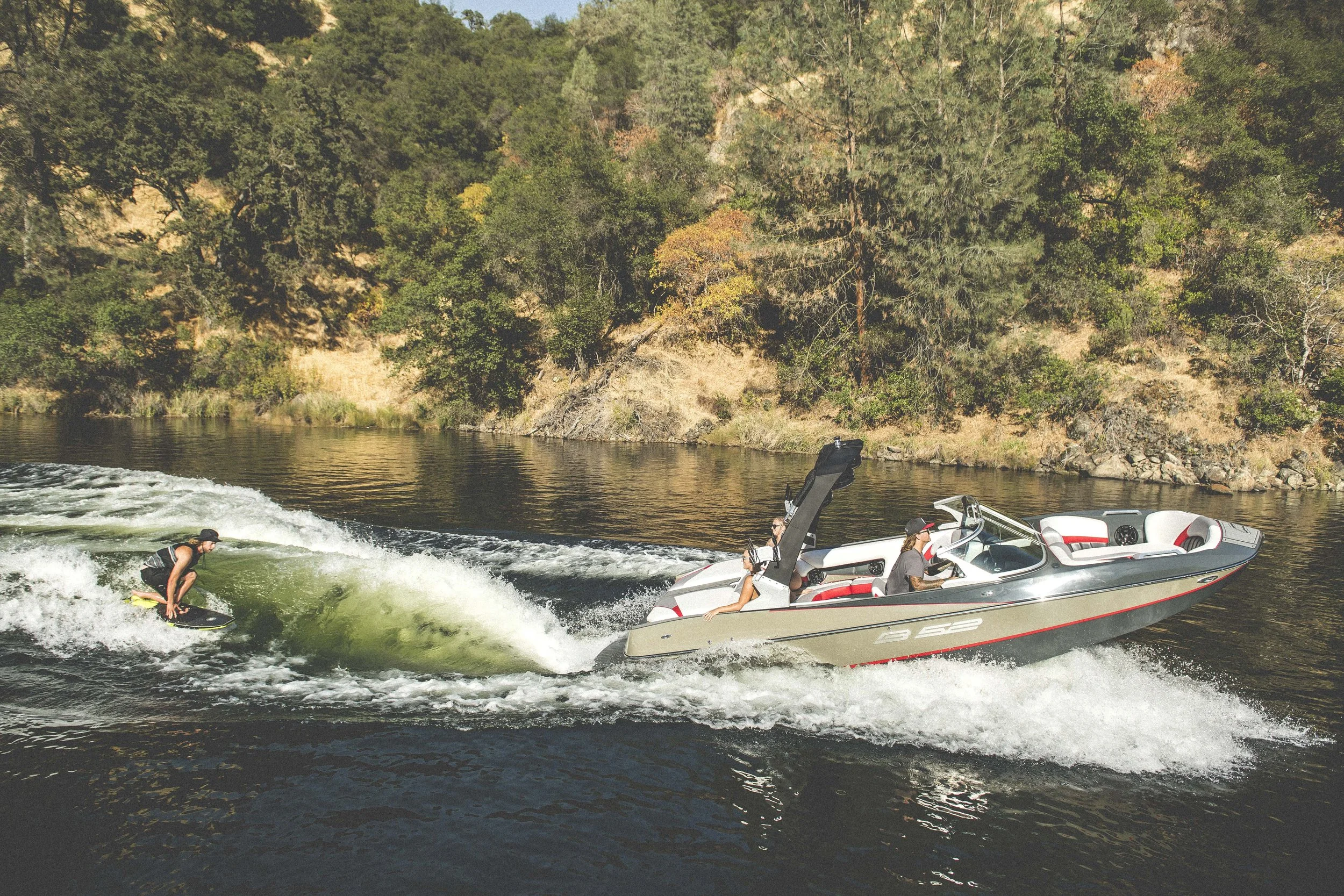 A person riding a wakeboard being pulled by a speedboat on a river with trees in the background.
