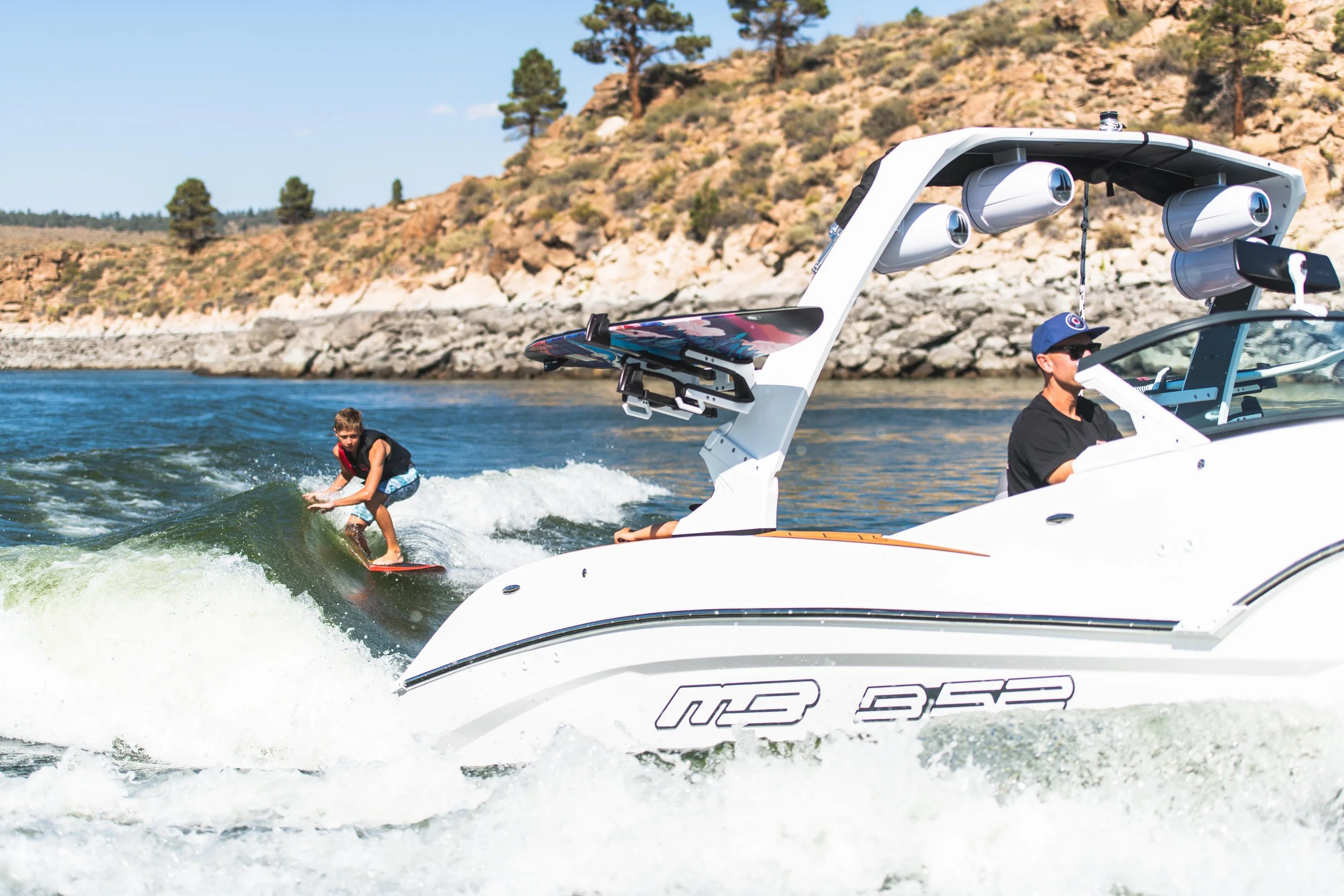 A boy riding a wakeboard behind a white speedboat on a lake with rocky shoreline and trees in the background.