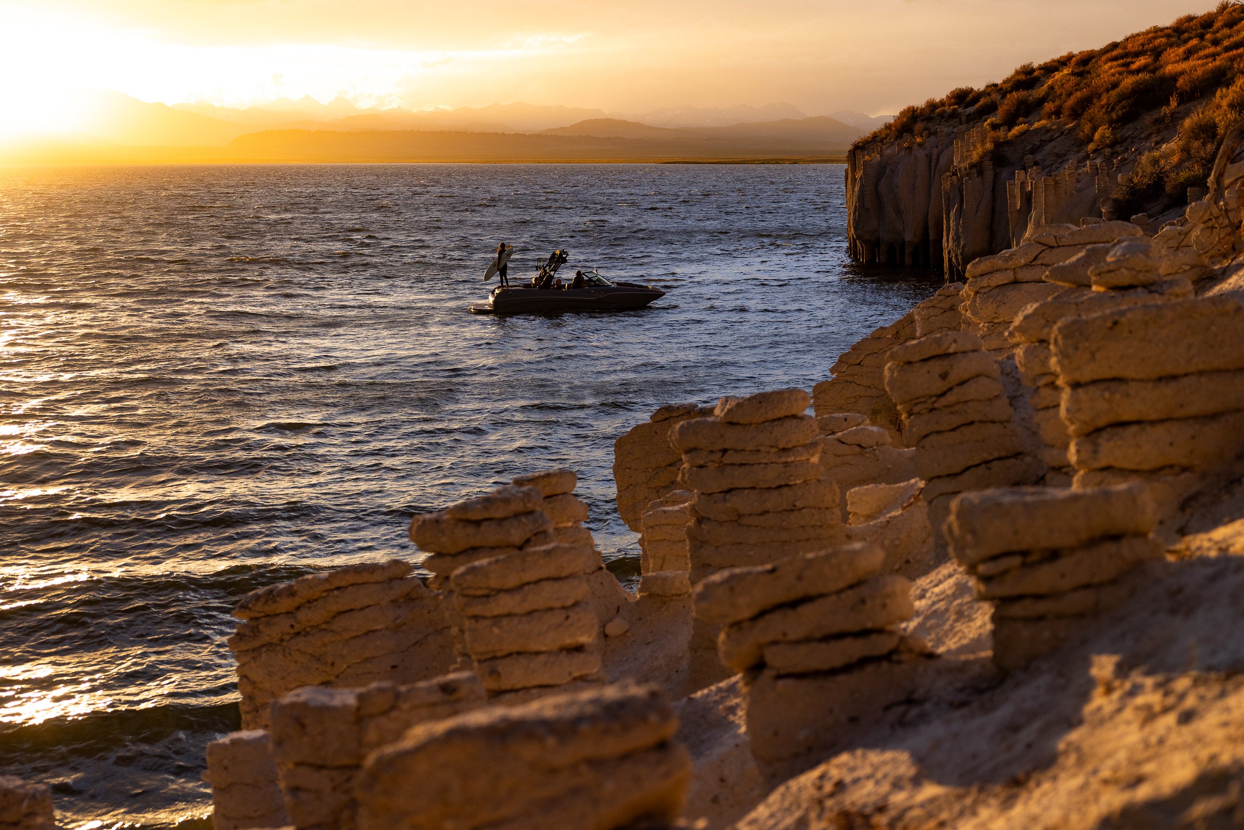 Sunset over a lake with a small boat and a person standing on the boat, near a rocky shoreline with layered rock formations.