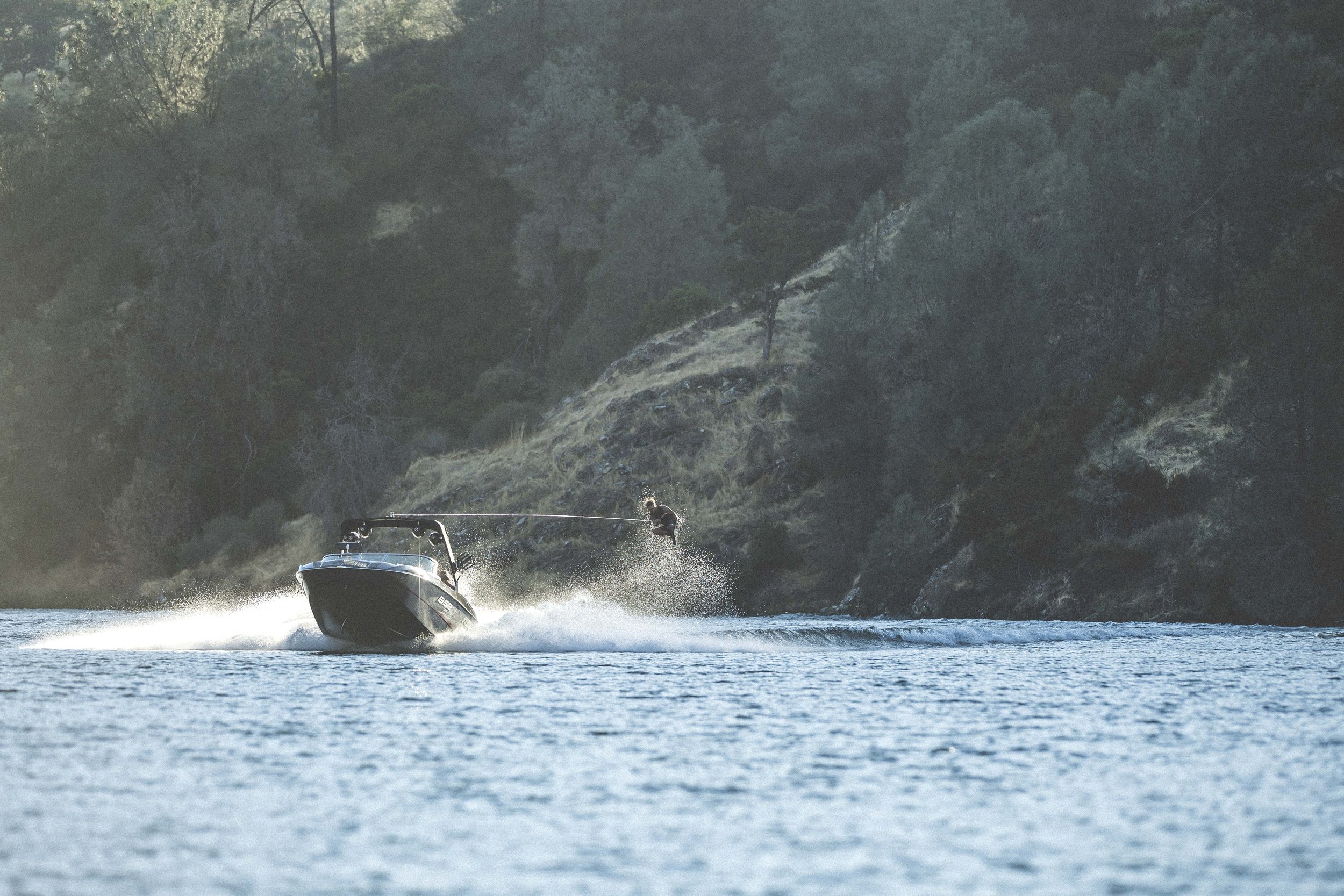A person wakeboarding on a large body of water near a densely wooded hillside, with the wakeboarder mid-air holding onto a tow rope.