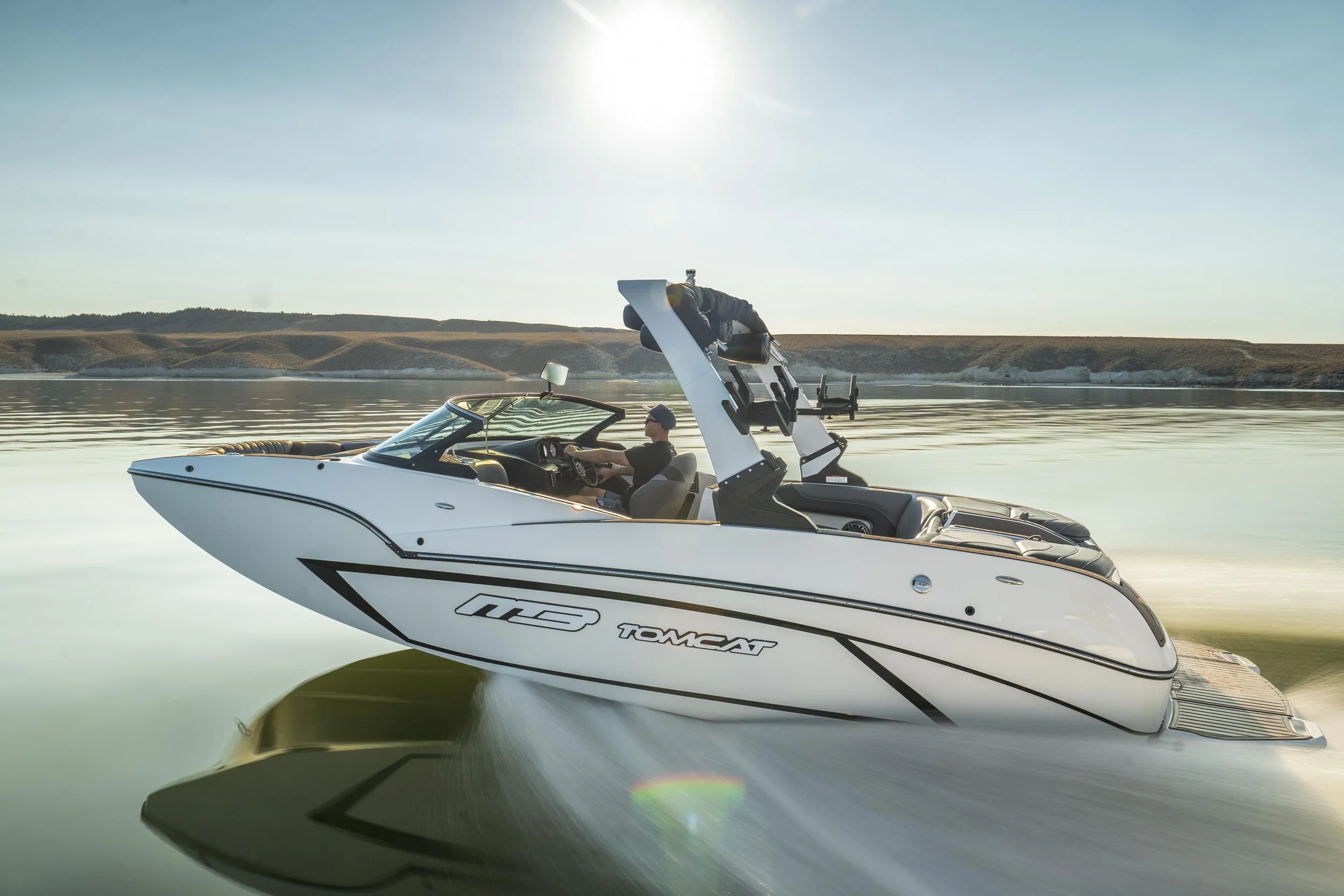 A white speedboat moving on calm water with a person at the helm and a man in a life jacket standing on the back, with a shoreline and hills in the background, under a clear sky.