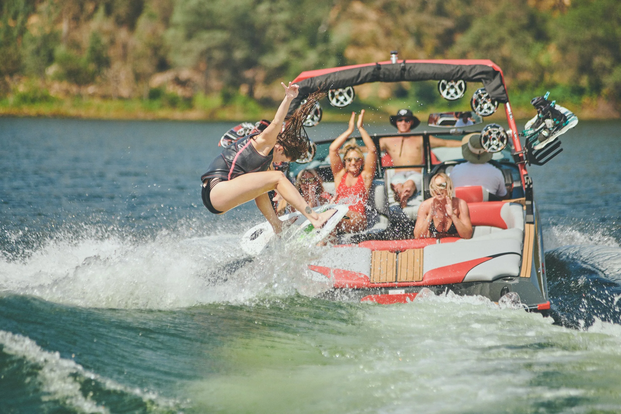 A person wearing a black life jacket is wakeboarding on a lake, with several people on a boat watching and enjoying the outdoor activity.