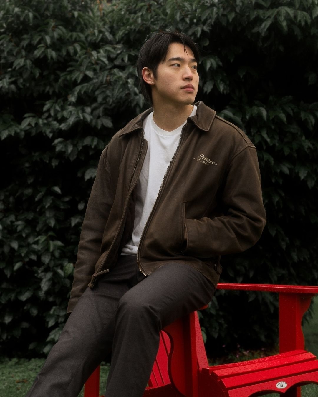 A young man sitting on a red park bench in Simon Fraser University Campus, Burnaby B.C.  with dark green bushes in the background, wearing a brown jacket and white t-shirt, looking to the side.