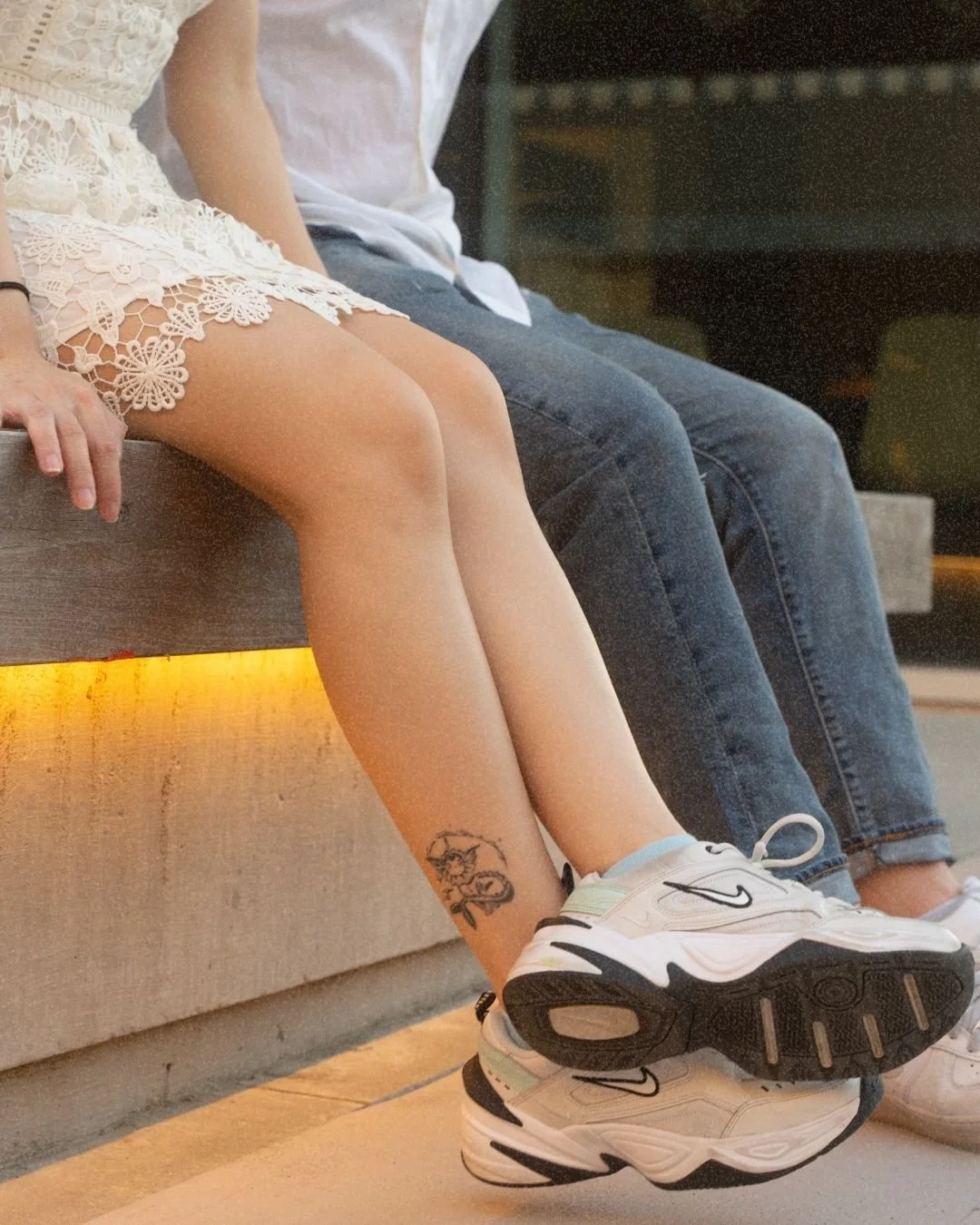 Close-up of two people sitting on a bench, showing only their legs and partly their torsos. One person is wearing white dress shoes and sports a tattoo on their lower leg. The other person is dressed in casual jeans. The seating area has warm ambient