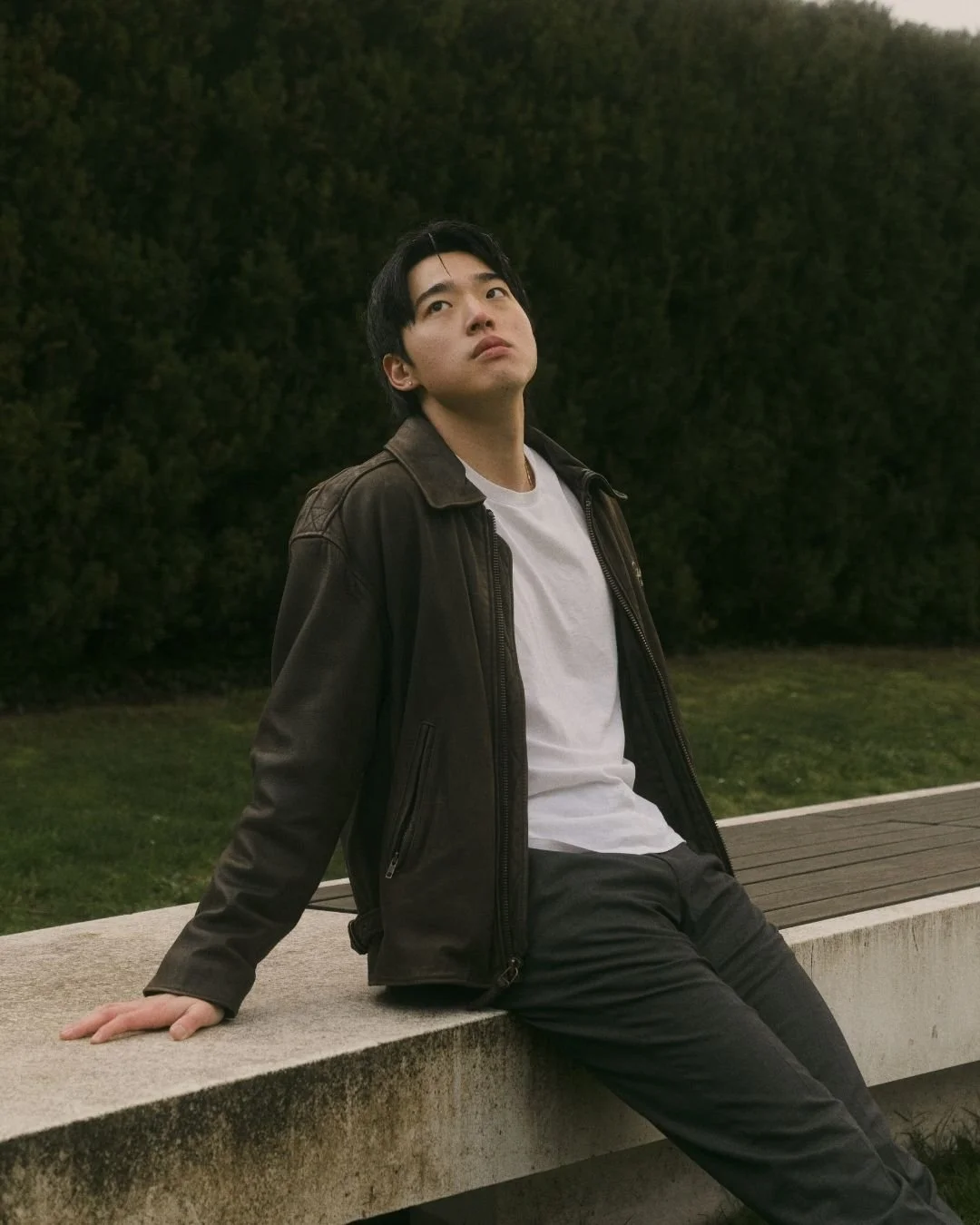 Young man sitting on a concrete ledge outdoors in Simon Fraser University Burnaby, B.C. looking up thoughtfully, with dark green bushes behind him.