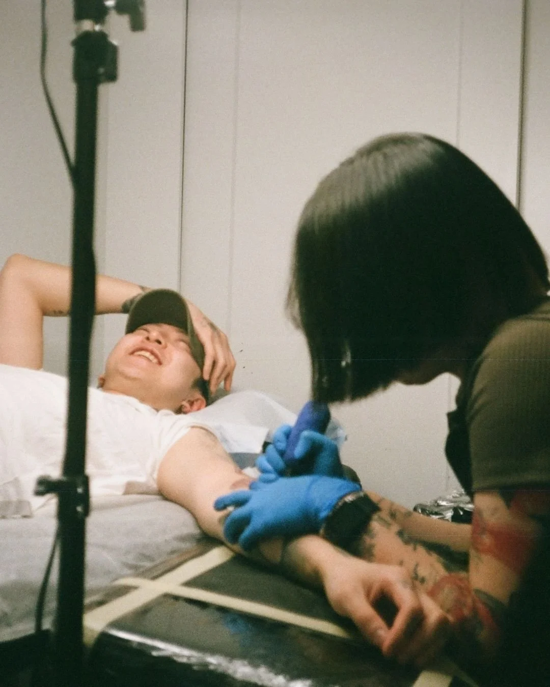 A person laying on a tattoo bed, smiling with a black cap, receiving a tattoo from a lady wearing blue gloves.