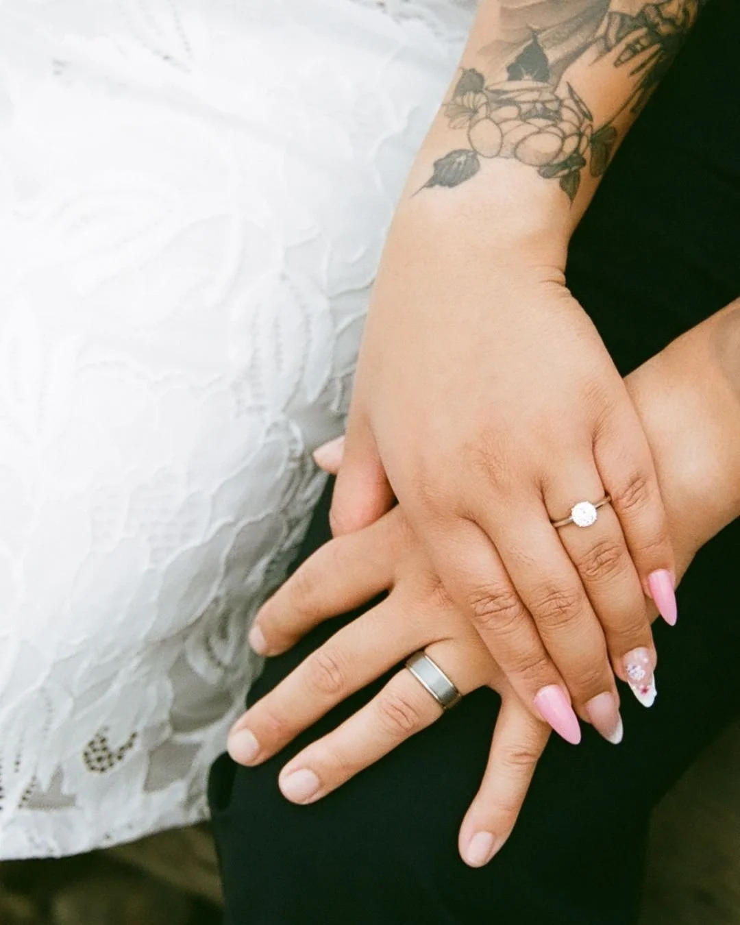 Close-up of two hands with wedding rings resting on each other. One hand has a diamond engagement ring, and the other a plain band. The person with the wedding ring has a tattoo on the wrist and hand. The background is a white textured surface.