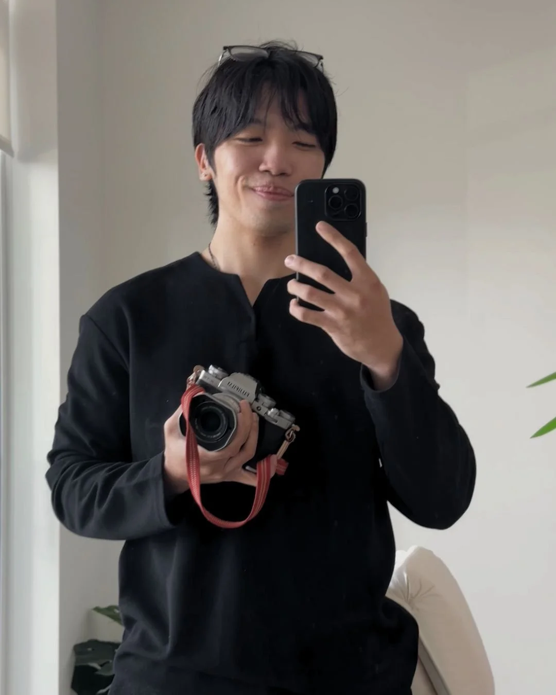 A young man smiling and taking a mirror selfie with a smartphone, holding a vintage camera in his left hand, wearing a black top and glasses on his head.