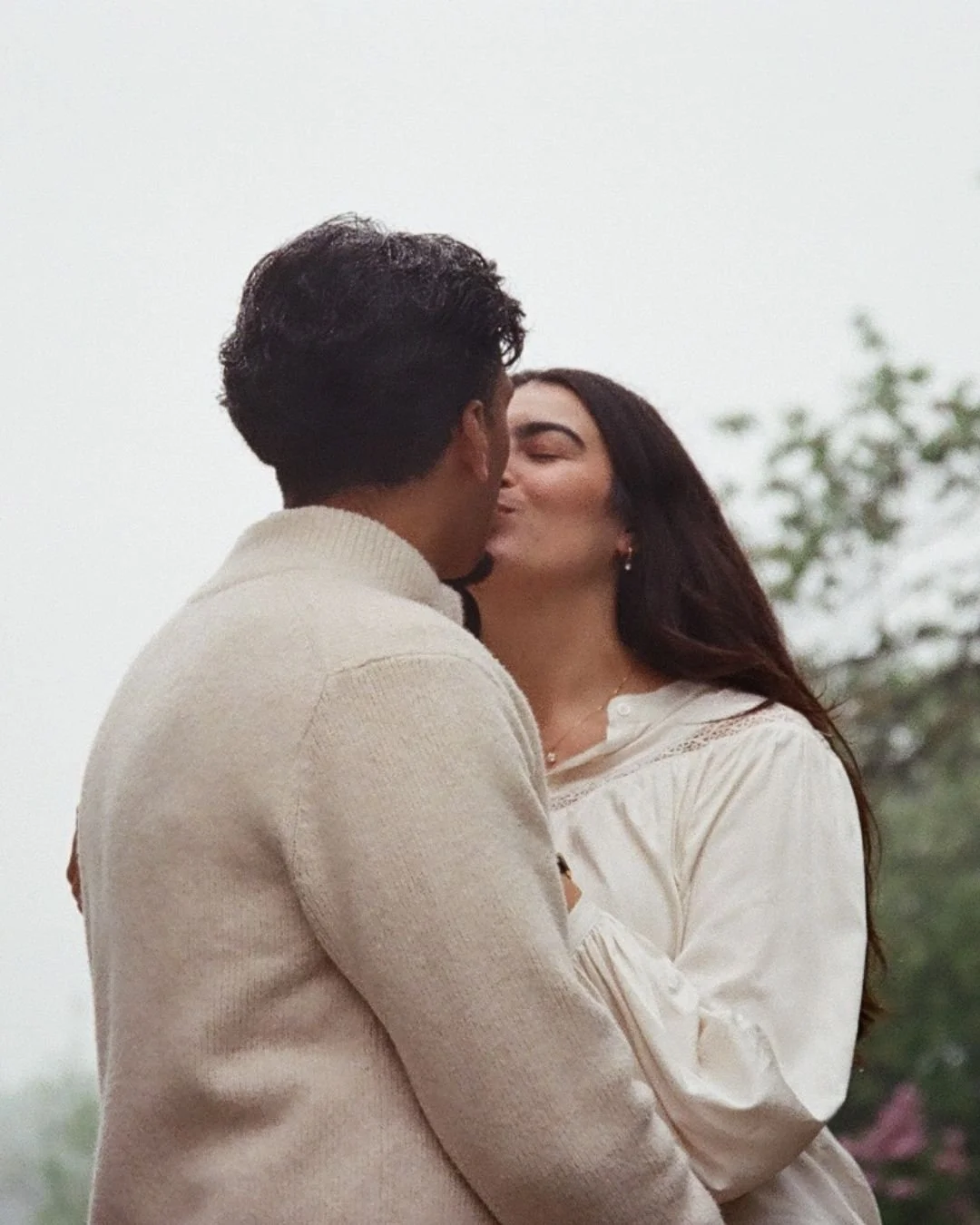 A man and woman share a kiss outdoors, with the man wearing a beige sweater and the woman in a white blouse, against a blurred background of trees and greenery.