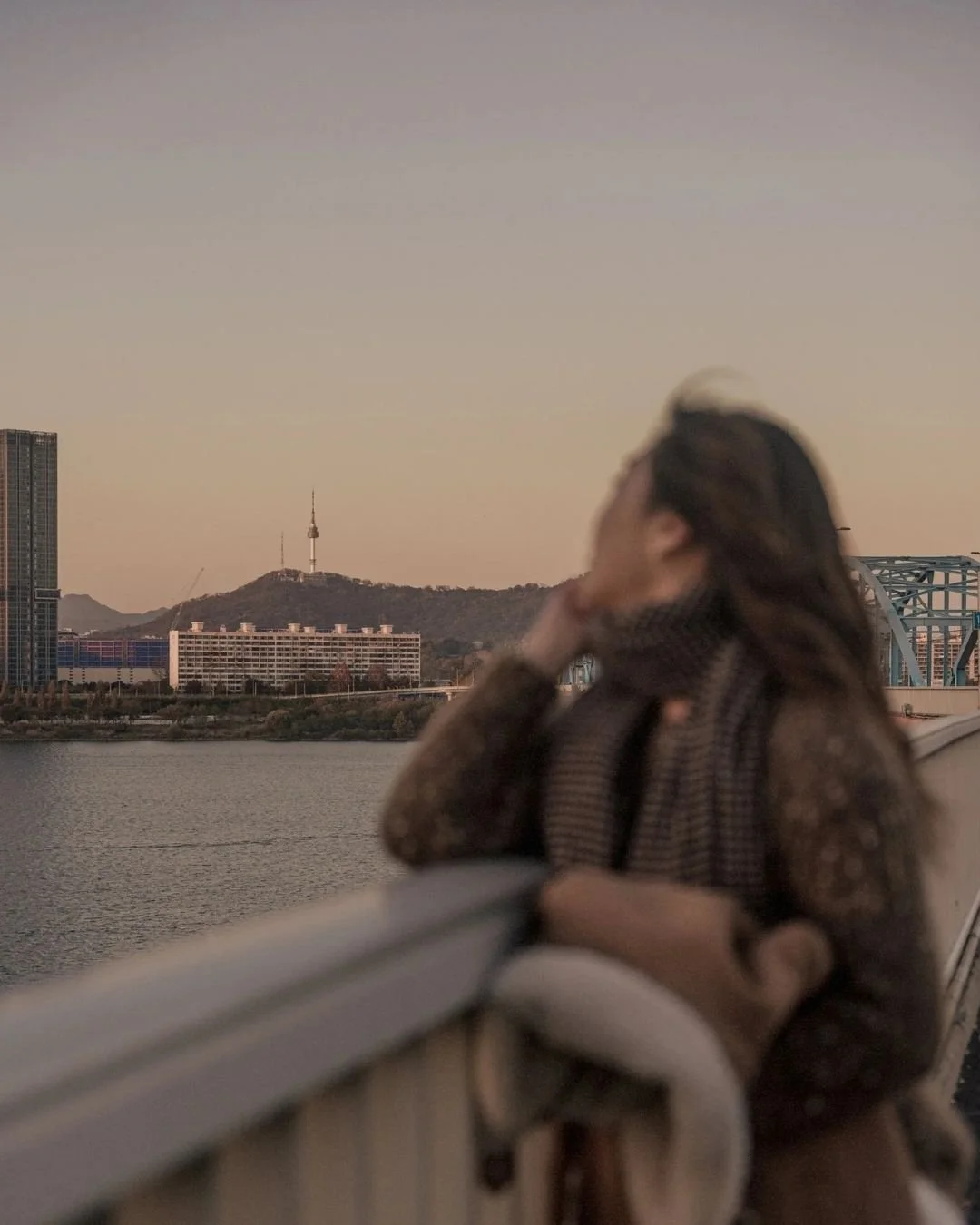A woman with long brown hair looking at a city skyline near a body of water during sunset.