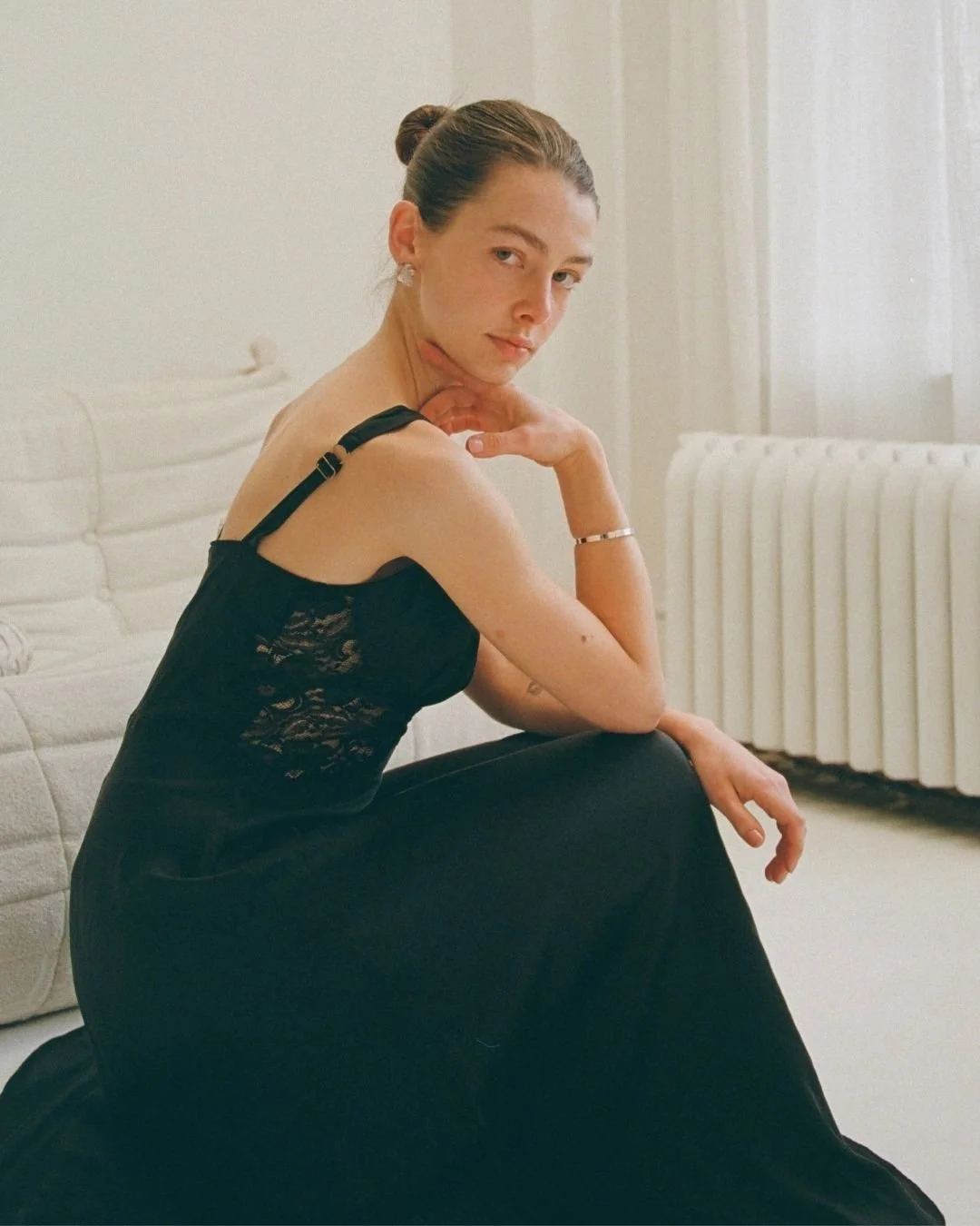 A woman with brown hair in a bun, wearing a black lace top and earrings, sitting on the floor in a room with white walls and furniture, resting her chin on her hand.