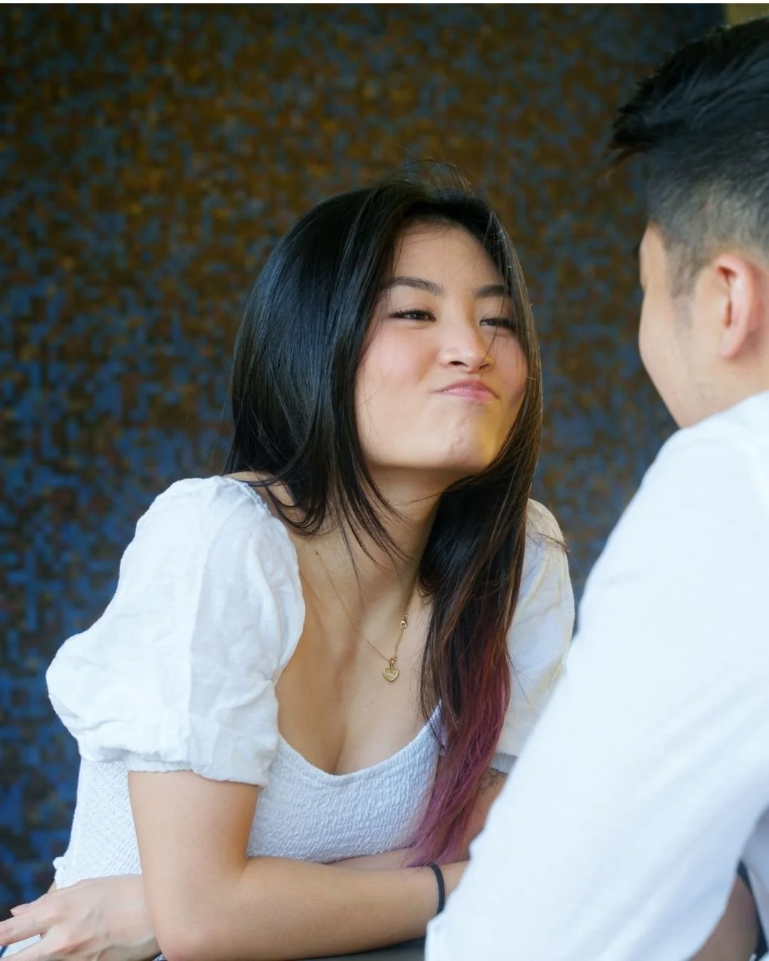 A young woman is making a playful facial expression while sitting across from a young man, both engaging in an interaction indoors with a textured dark background. In Burnaby B.C.