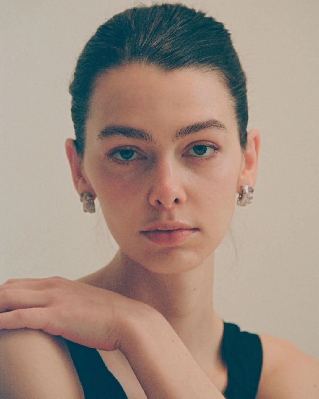 Close-up photo of a woman with dark brown hair pulled back, wearing earrings and a black sleeveless top, gazing at the camera with a neutral expression, against a plain background.