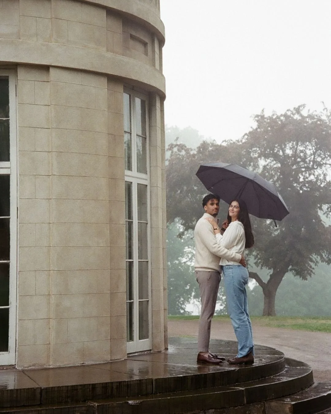 A couple standing on a rain-soaked platform under a black umbrella next to a beige stone building with large windows, with trees and foggy weather in the background.