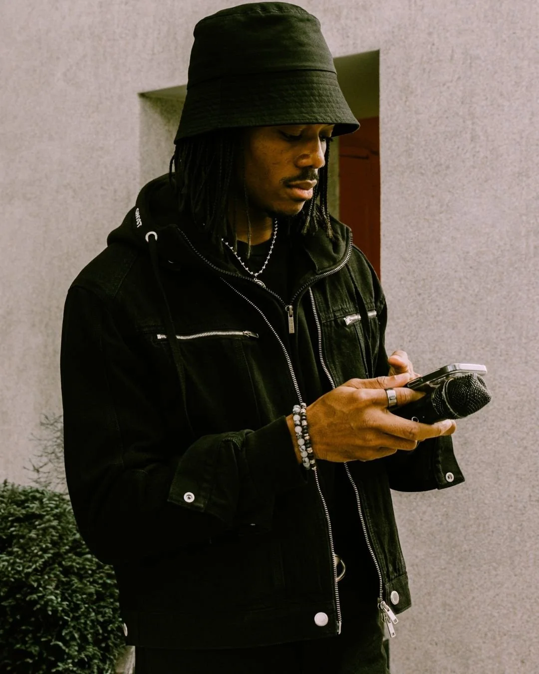 Young man with dreadlocks wearing a black bucket hat and black jacket, looking at his phone, standing against a grey wall.