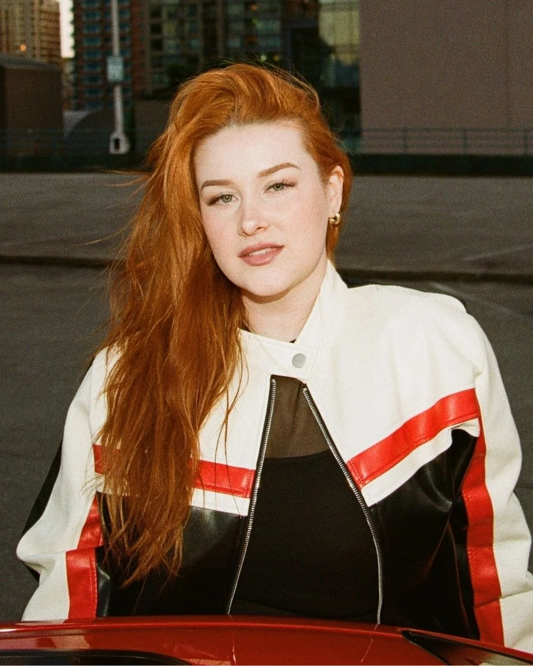 A woman with long red hair, wearing a white jacket with red and black accents, sitting outdoors with a cityscape in the background.