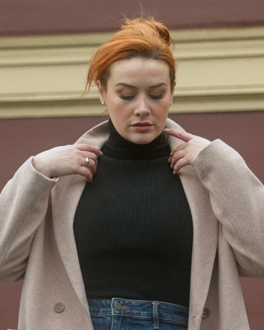 A woman with red hair, wearing a black turtleneck and a beige coat, stands with her eyes closed and hands adjusting her collar, against a plain wall background.