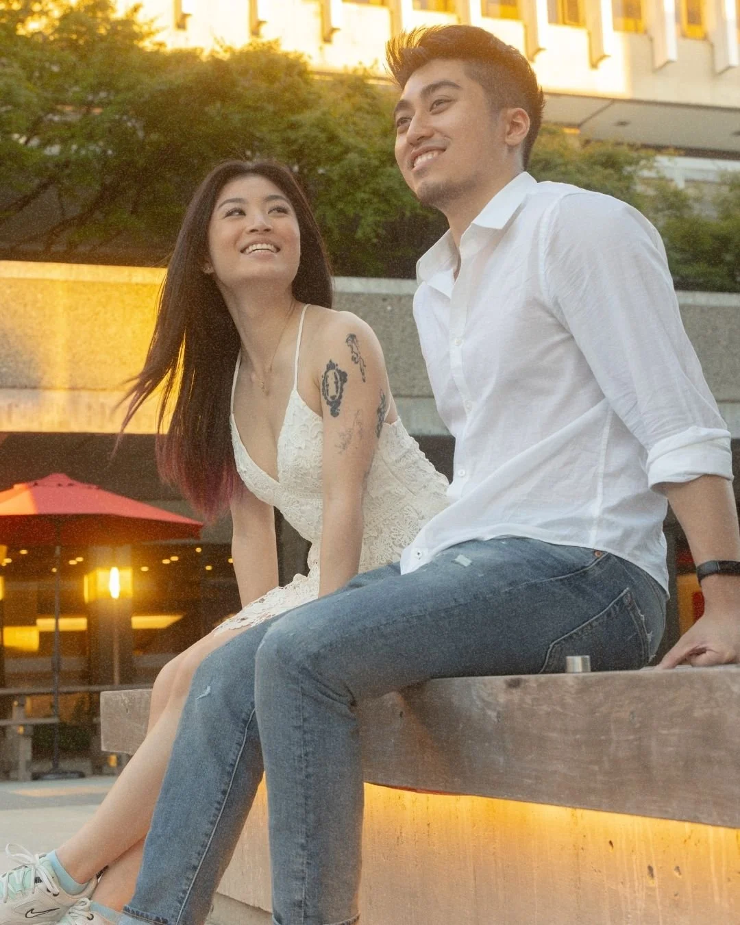 A young Asian woman and a young Asian man sitting on a concrete bench outdoors during sunset, smiling and looking at each other. She is wearing a white lace dress and has tattoos on her arm, while he is wearing a white button-up shirt and jeans.