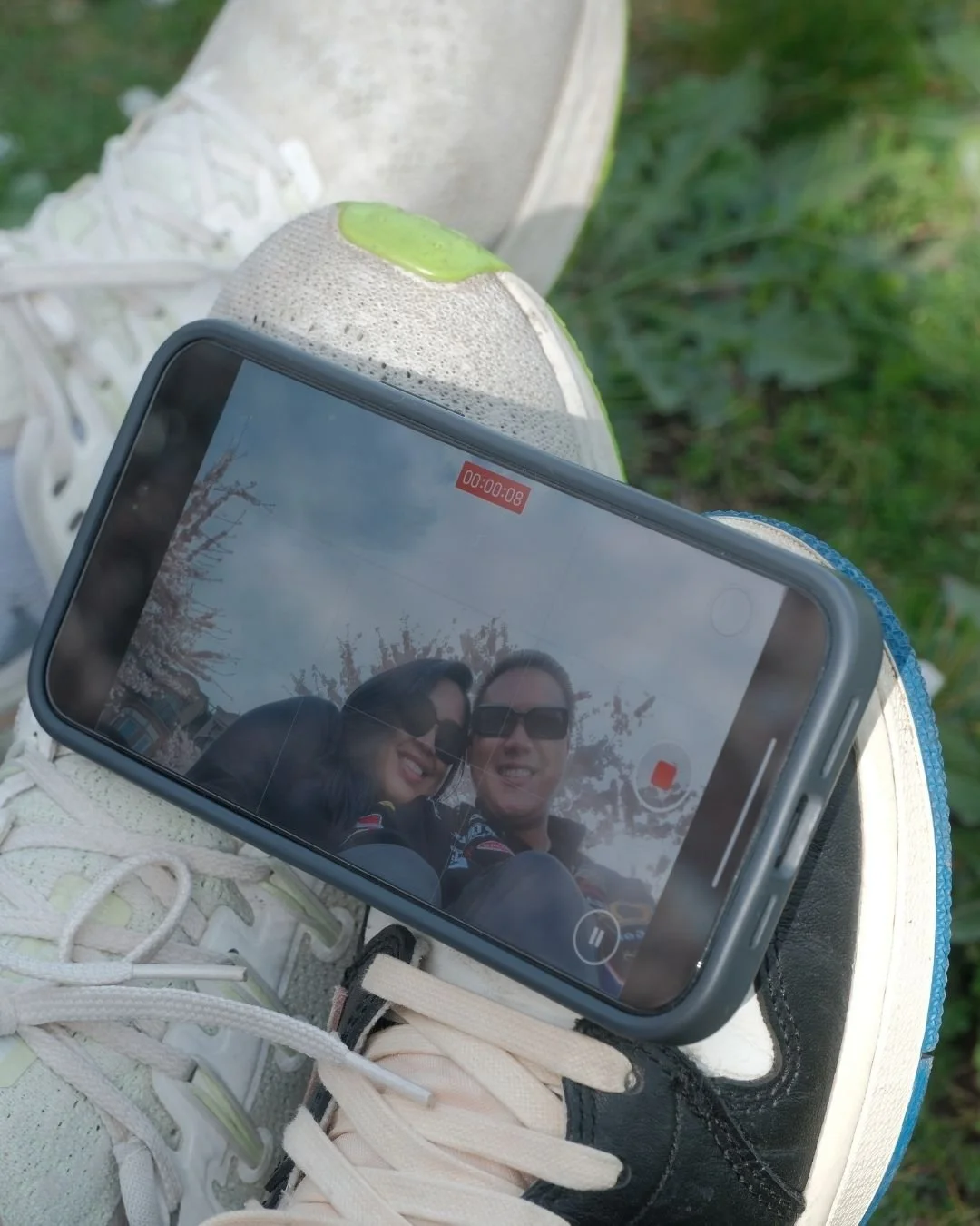 A smartphone resting on white sneakers with green and black accents, capturing a selfie of a smiling couple outdoors, with trees and pink blossoms reflected on the phone screen.