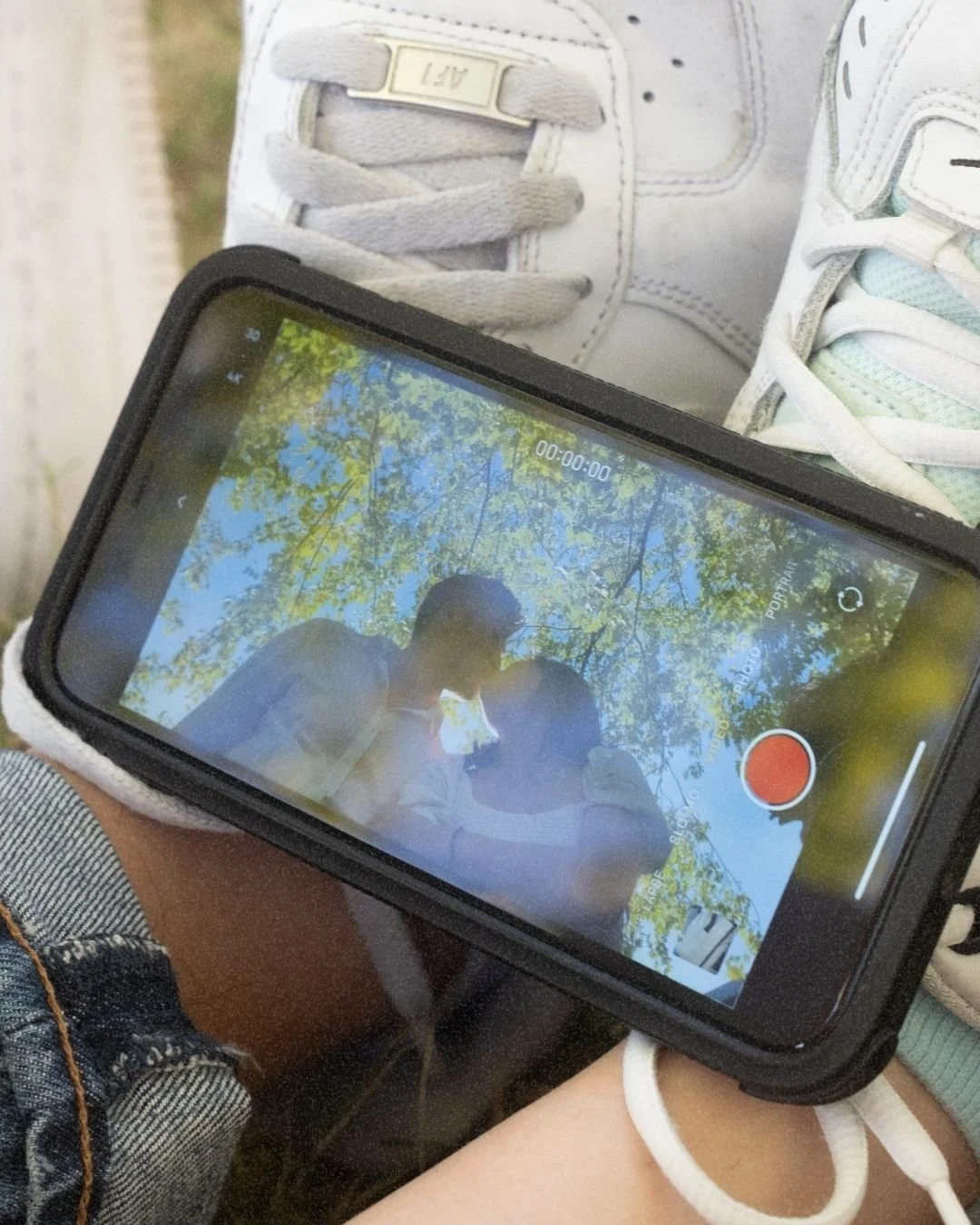 A person holding a smartphone shows a recorded video of two people kissing outdoors under tree branches with green foliage.