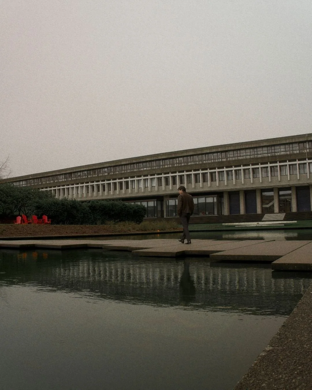 A man walking on a floating platform over water in front of a modern, multi-story building with large windows and a flat roof, on a cloudy day.